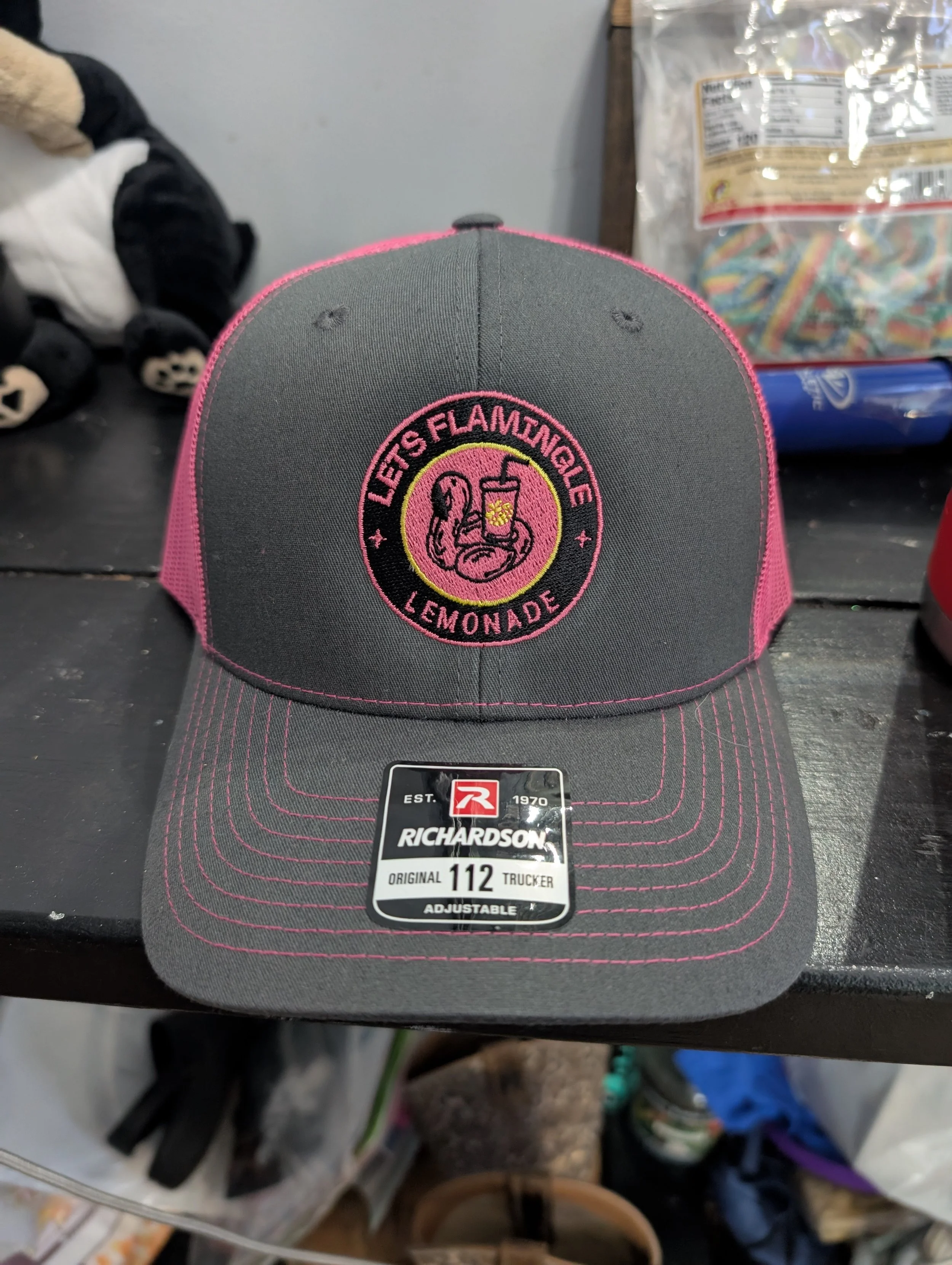 Gray and pink trucker cap with embroidered patch reading 'Let's Flamingo Lemonade' featuring a flamingo drinking from a lemonade glass, placed on a black surface.
