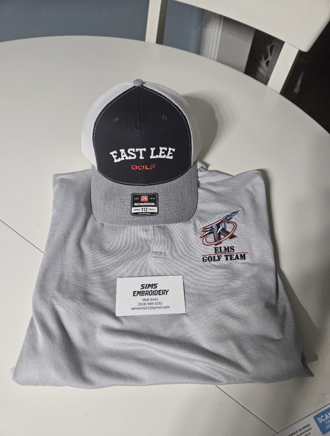 A gray and black East Lee Golf cap resting on a gray embroidered ELMS Golf Team polo shirt on a white surface.