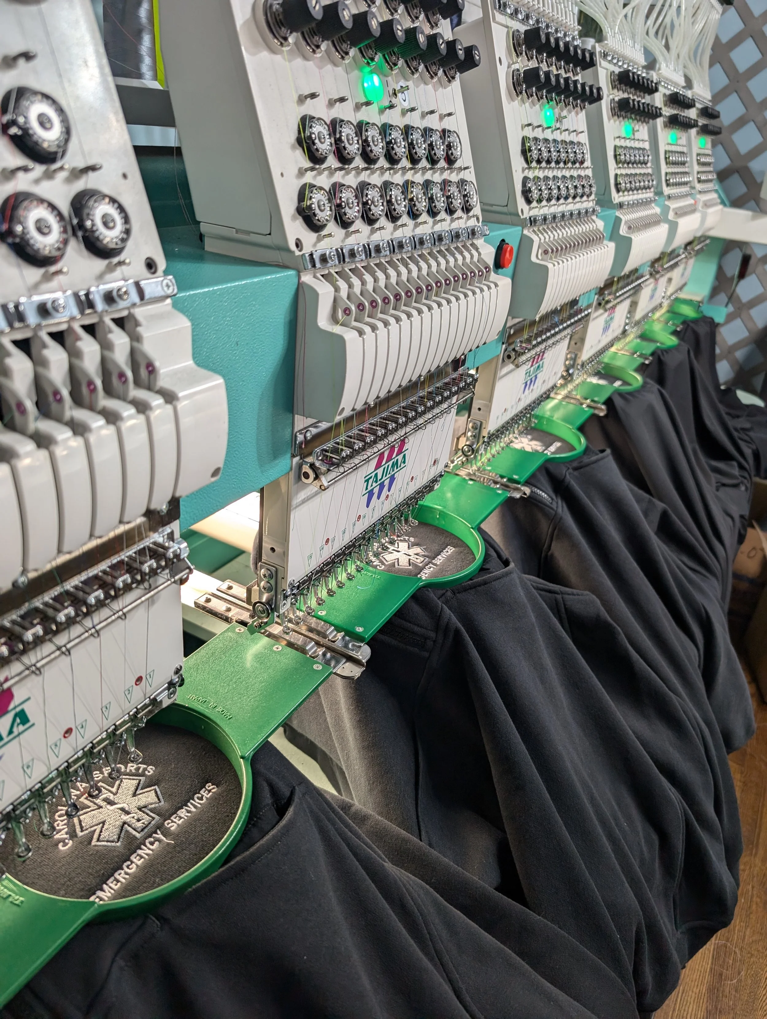 Close-up view of multiple embroidery machines stitching black fabric with patches that display medical symbols and text, set in an organized workshop.