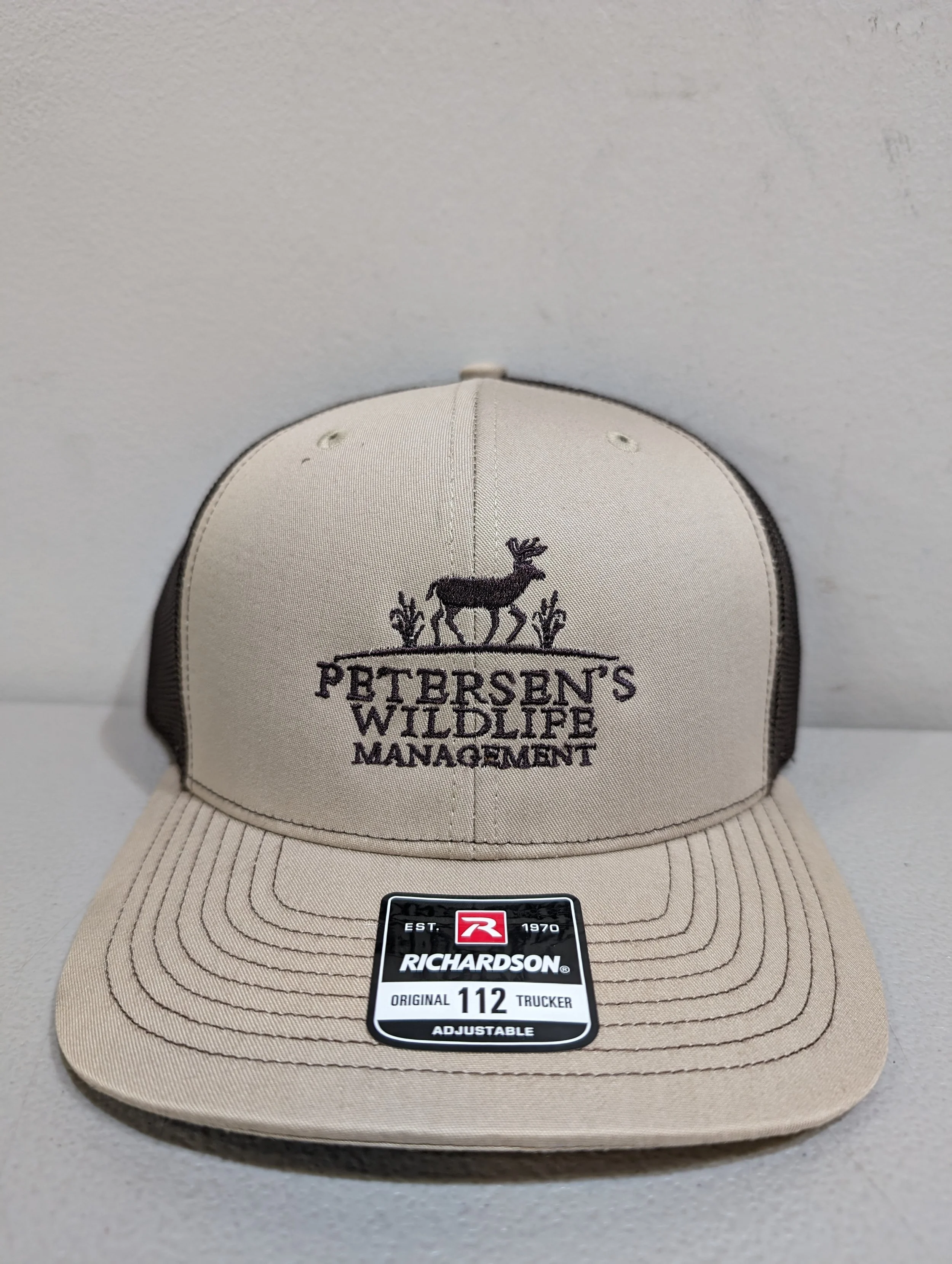 Beige trucker cap with black mesh back, embroidery of a deer and plants, and text 'Petersen's Wildlife Management'.