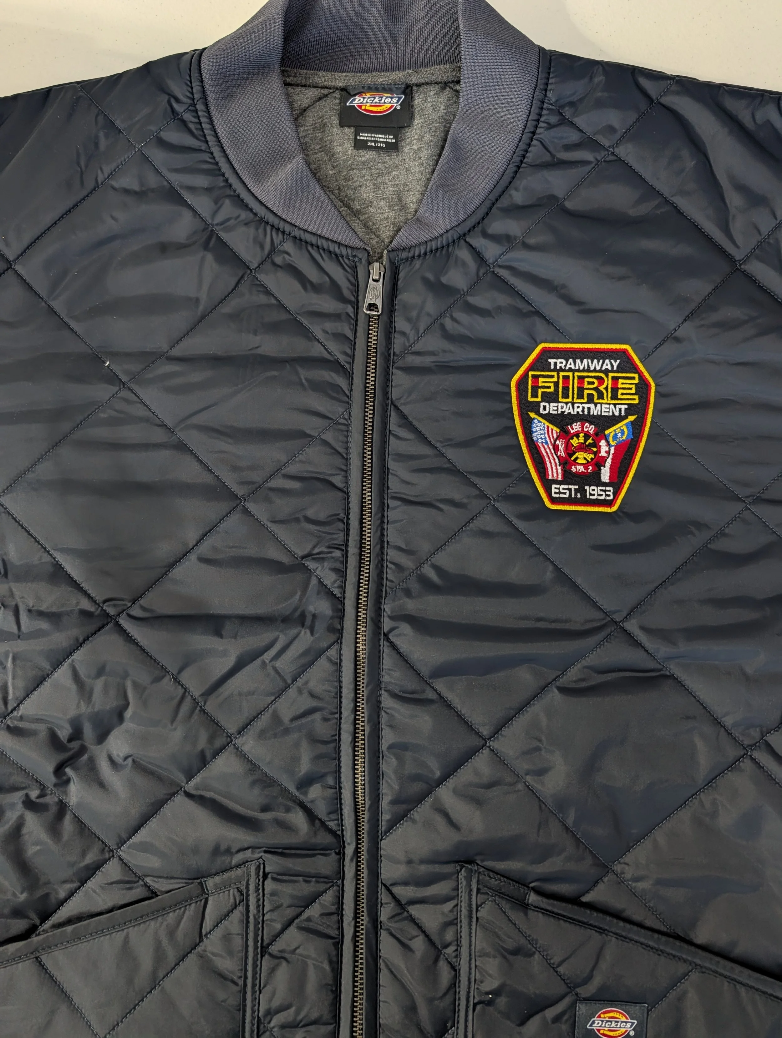 Black quilted jacket with a badge on the chest reading 'Tramway Fire Department' and featuring various patches, a zipper in the center, and a Dickies label inside the collar.