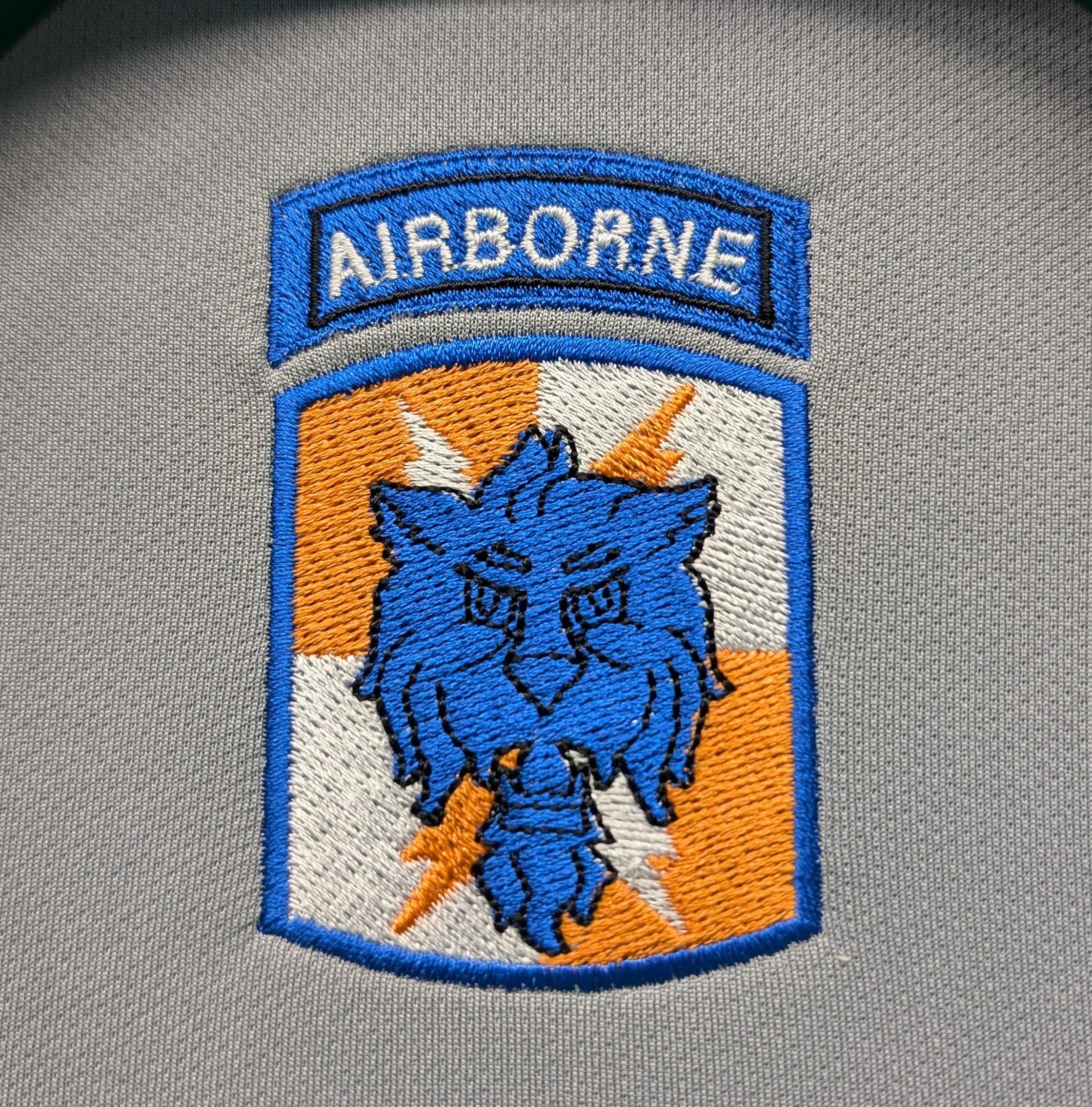 A patched emblem with the word 'AIRBORNE' at the top, featuring a blue lion's head with a fierce expression, lightning bolts in the background, and an orange and white striped background.