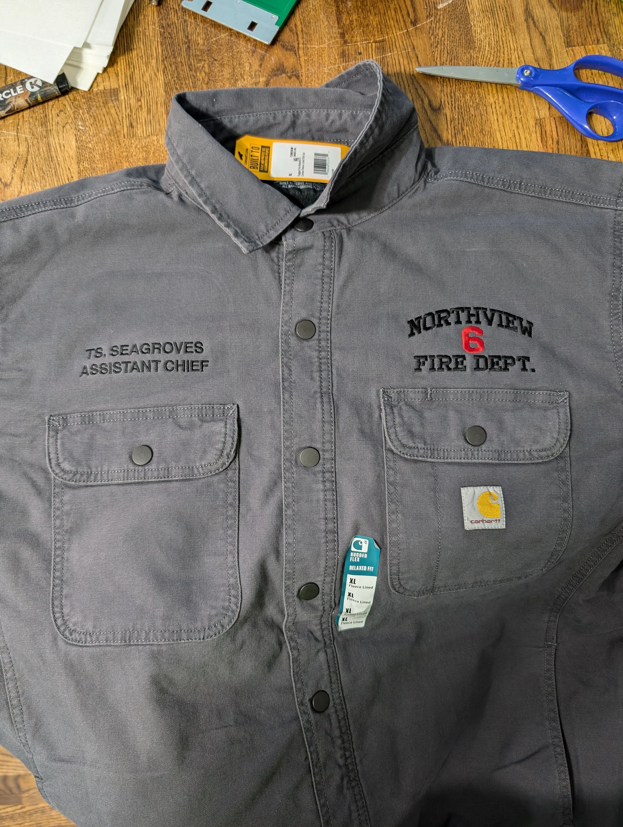 Gray uniform shirt with fire department patches, including 'T.S. Seagroves Assistant Chief' and 'Northview Fire Dept.' with a red number 6, on a wooden table with scissors and a tag nearby.