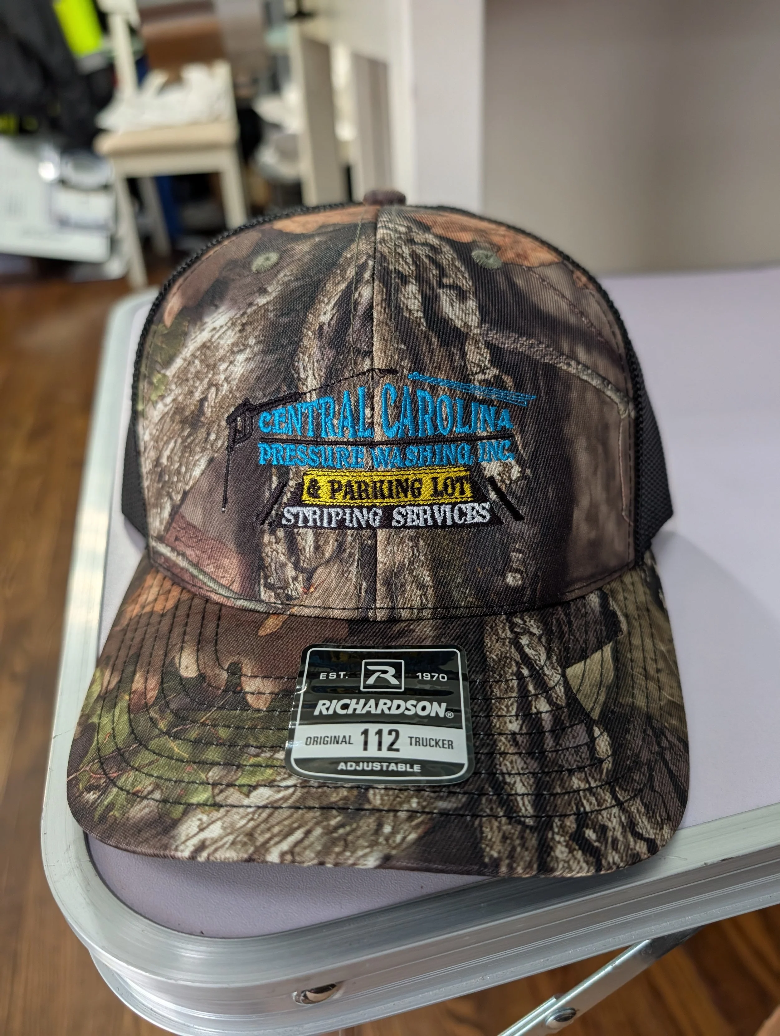 Camouflage baseball cap with embroidered logo of Central Carolina Pressure Washing Inc. and striping services, on a purple surface.