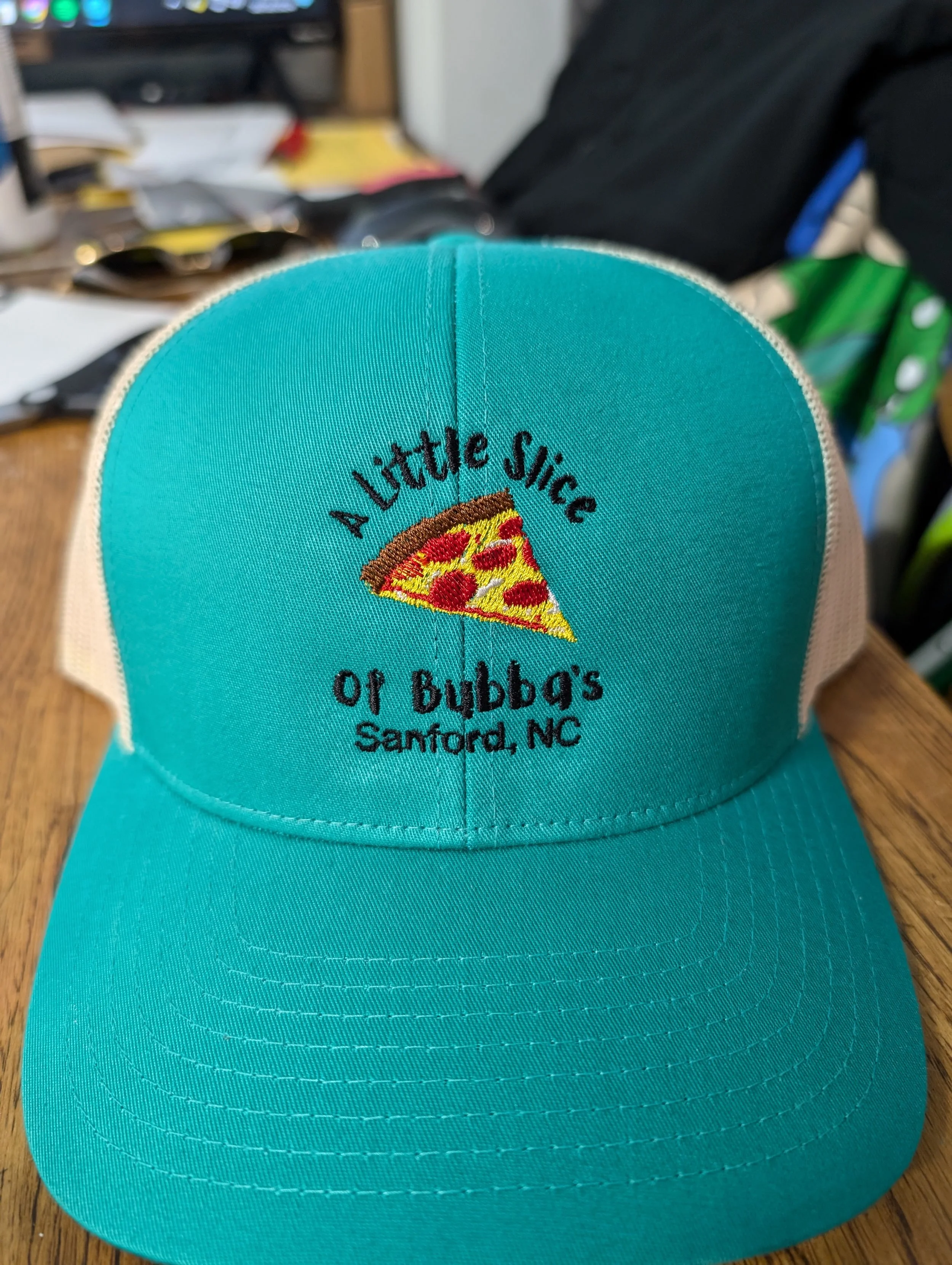 A turquoise and white trucker hat with an embroidered pizza slice and the words "A Little Slice of Bubba's Sanford, NC" on the front