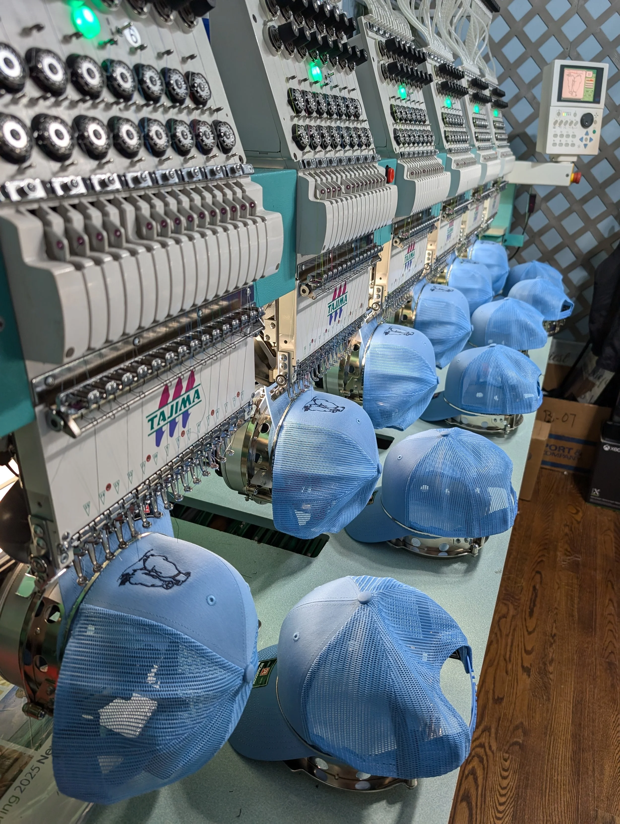 Multiple embroidery machines sewing light blue baseball caps with mesh backs on a green work surface.