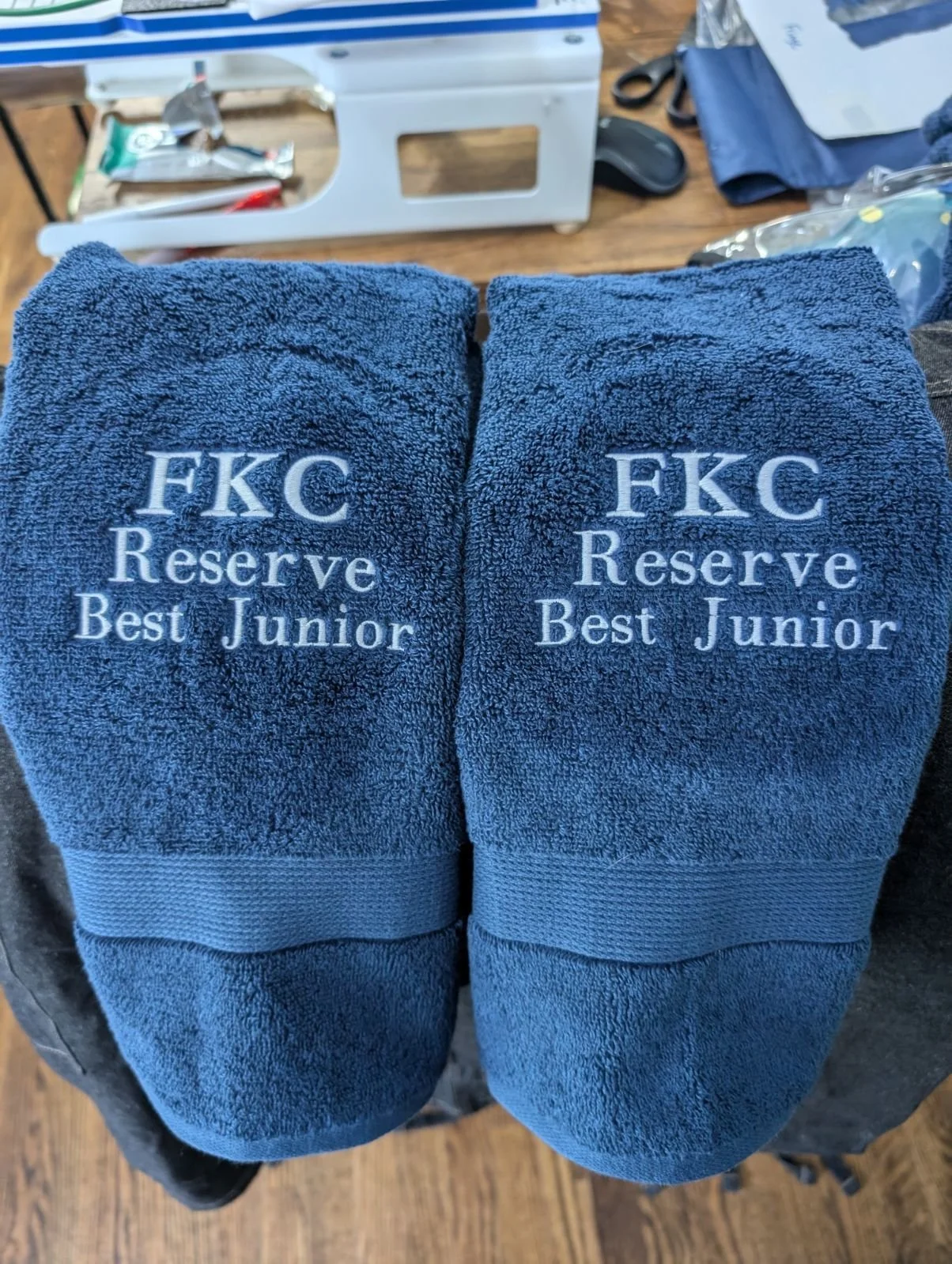 Pair of blue towels embroidered with 'FKC Reserve Best Junior' placed on a dark table, with a workspace in the background.