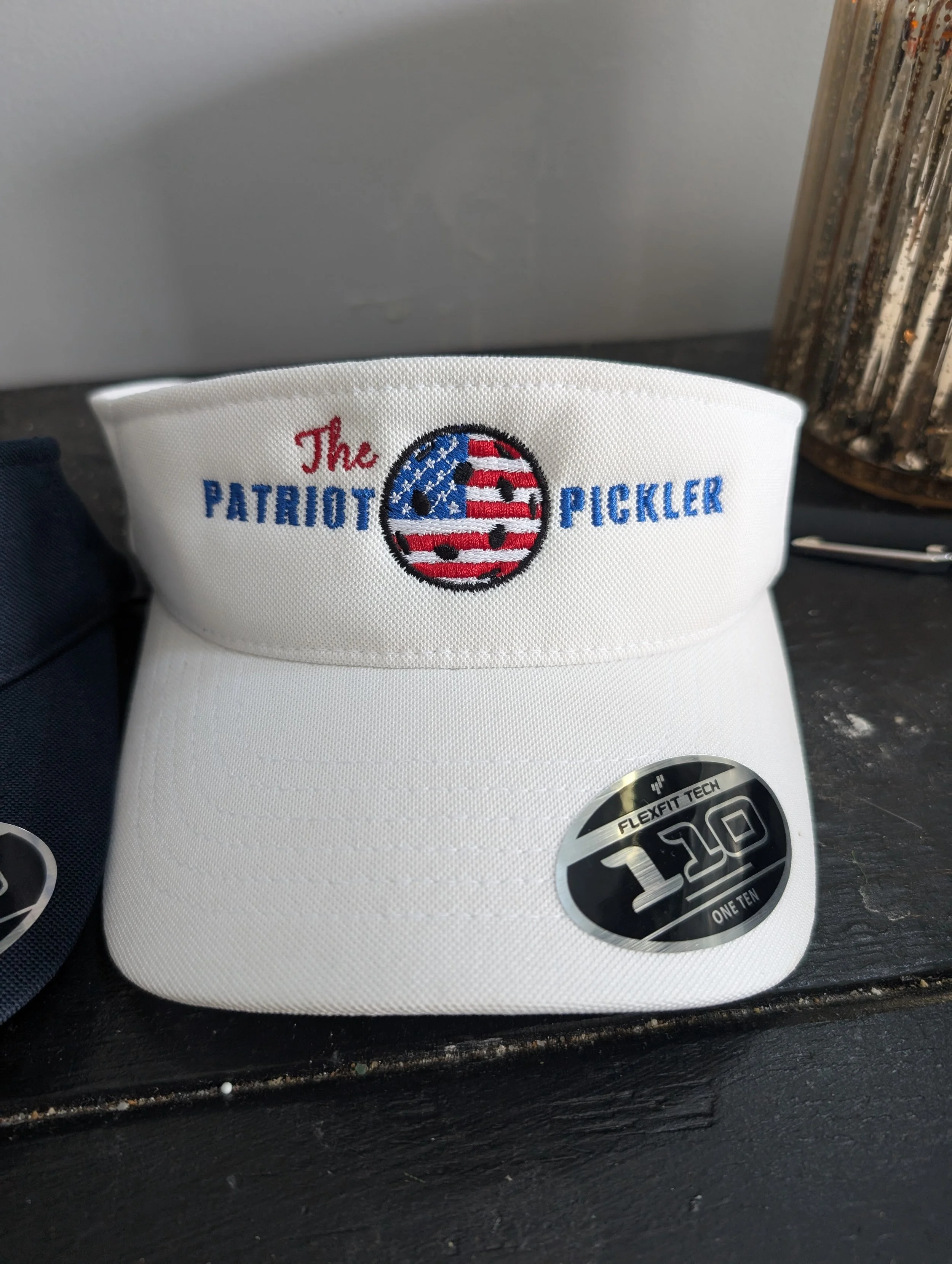 A white sports visor cap with embroidery that reads 'The Patriot Pickler' and features a basketball with an American flag design. There is a sticker on the brim indicating 'Flexfit Tech 110 ONE TEN'.