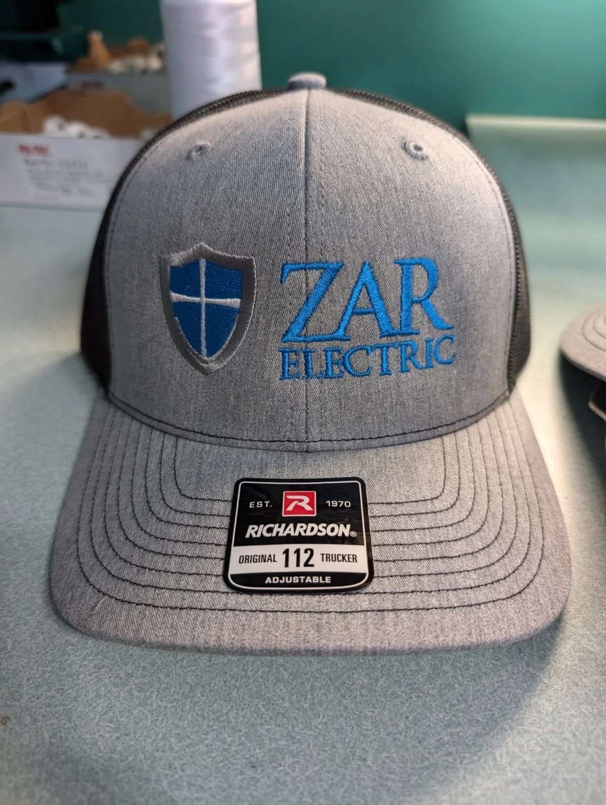 Gray baseball cap with embroidered blue shield and text 'ZAR Electric', displayed on a table.