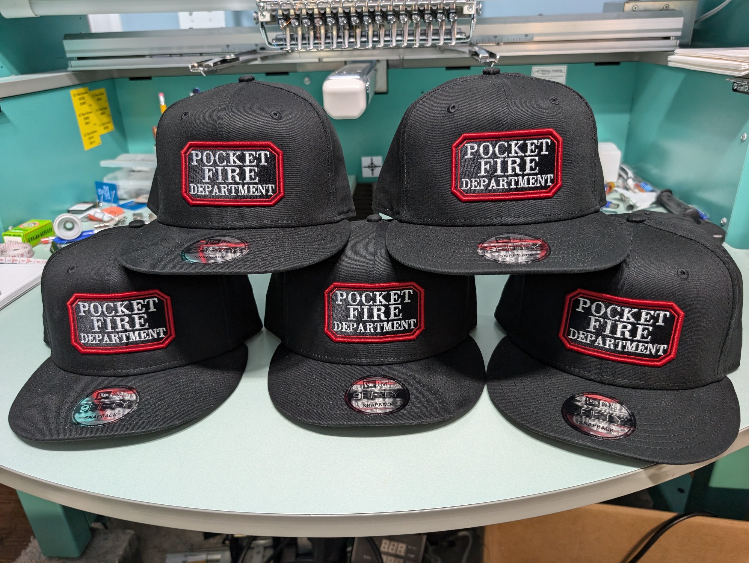 Six black hats with embroidered patches that say 'Pocket Fire Department' arranged on a table in a workshop or store