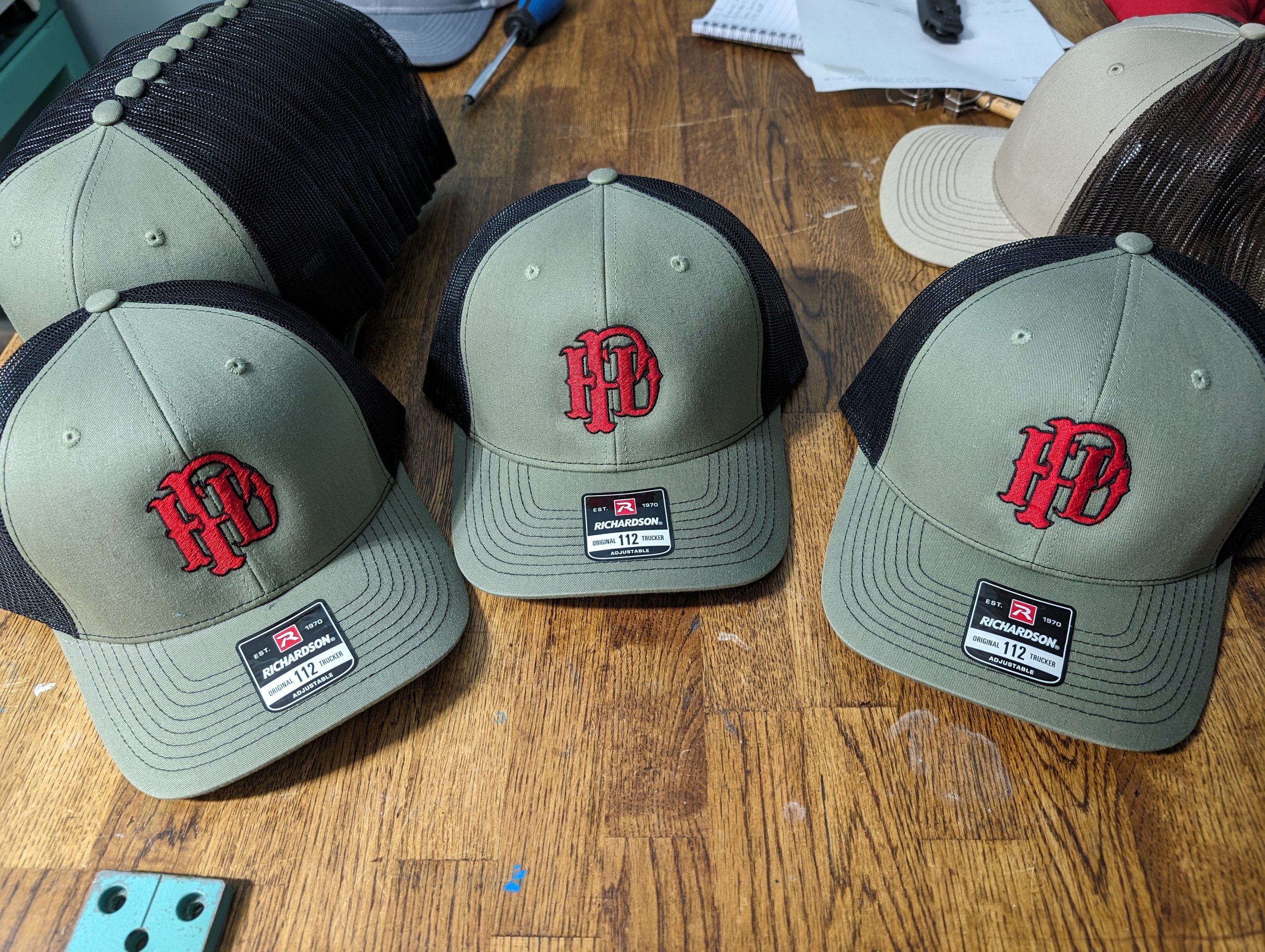 A display of olive green and black mesh trucker hats with red embroidered initials on the front, placed on a wooden table.