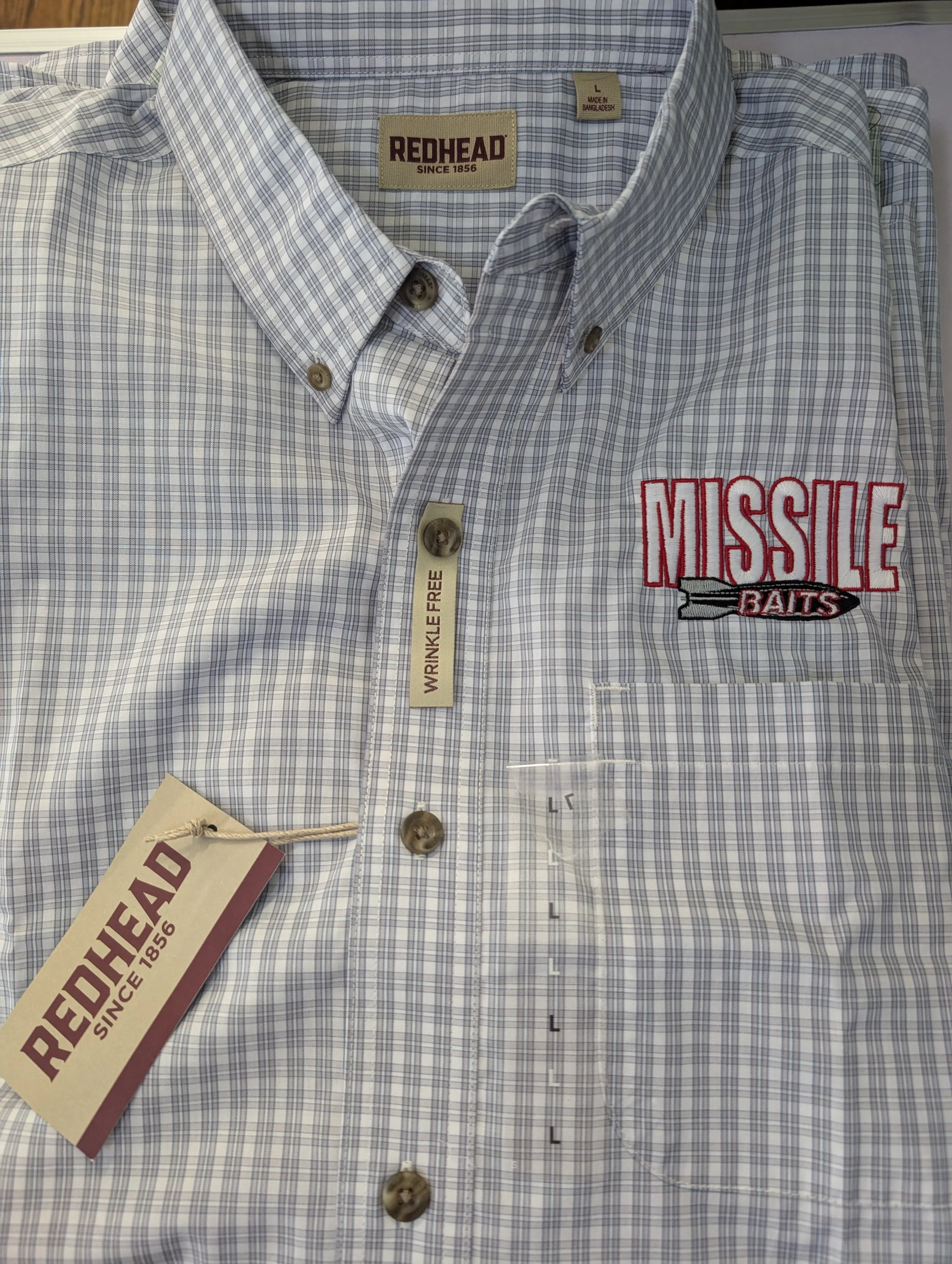 Close-up of a men's plaid button-up shirt with an embroidered patch reading 'MISSILE BAITS' and tags indicating 'RedHead' brand, size large, made in Bangladesh, wrinkle-free.