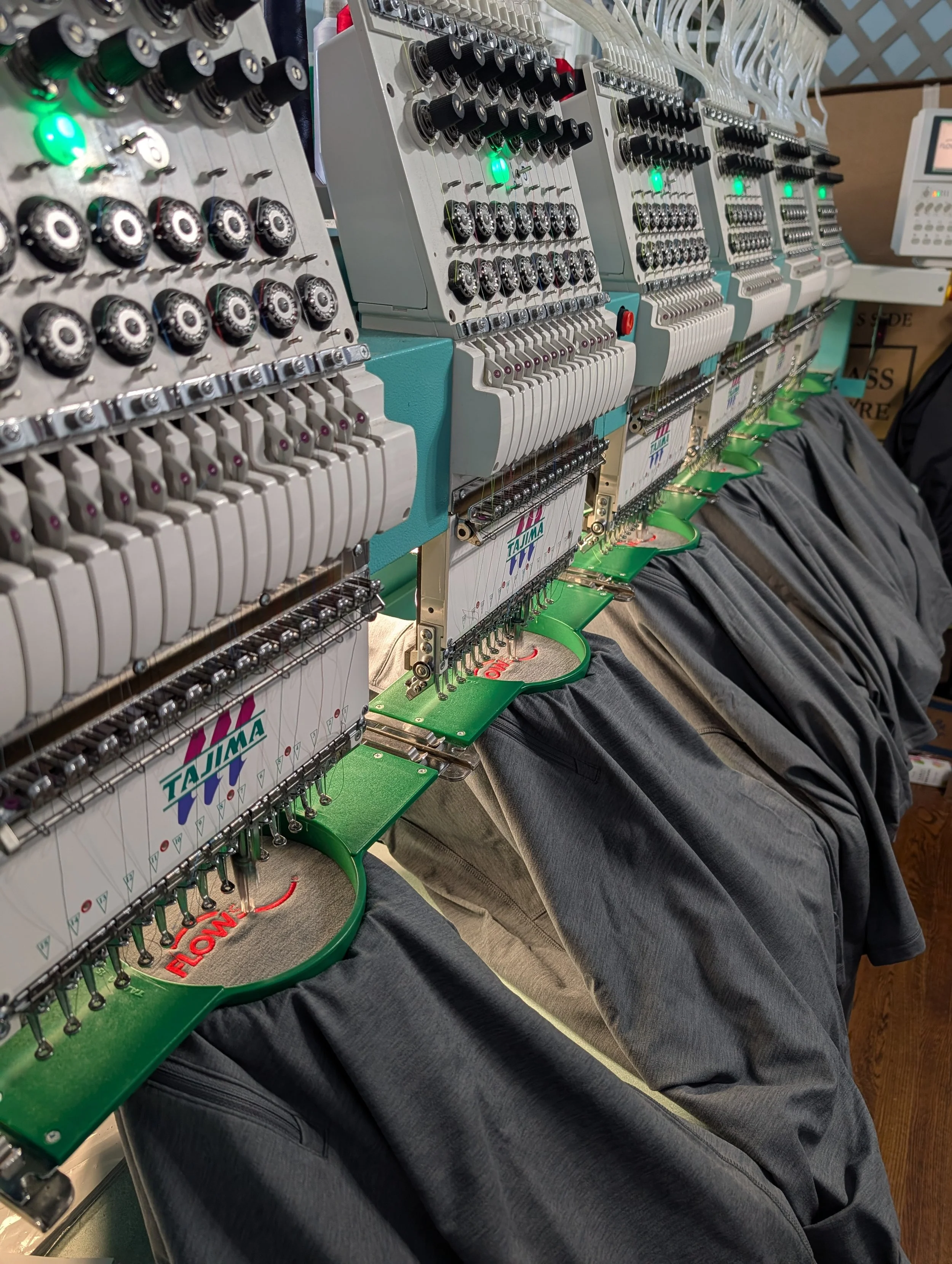 Close-up of multiple embroidery machines sewing fabric with the brand name 'TAJIMA' visible on the machine heads.
