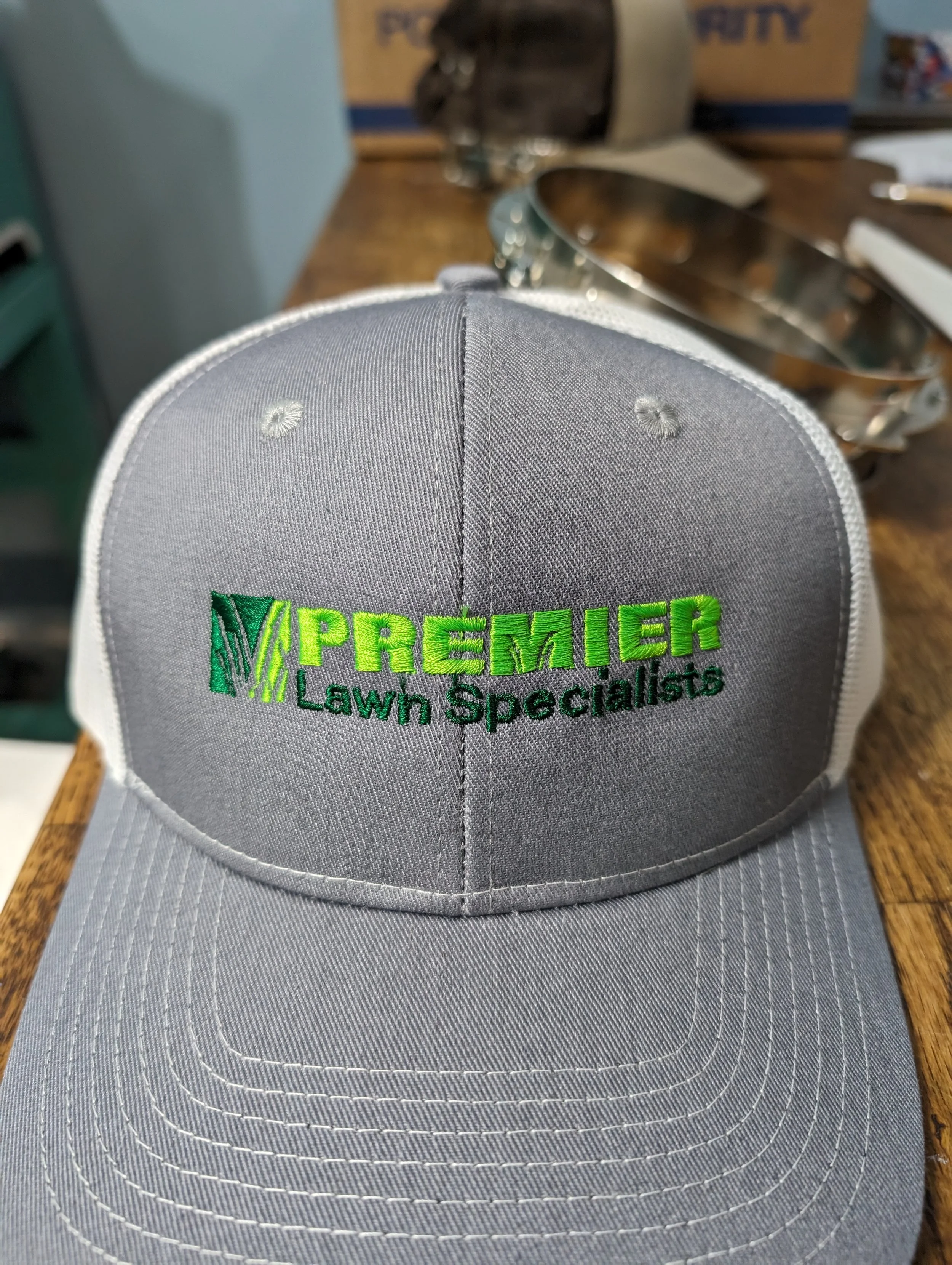 Grey and white baseball cap embroidered with green and black text reading 'PREMIER Lawn Specialists' on a wooden surface.