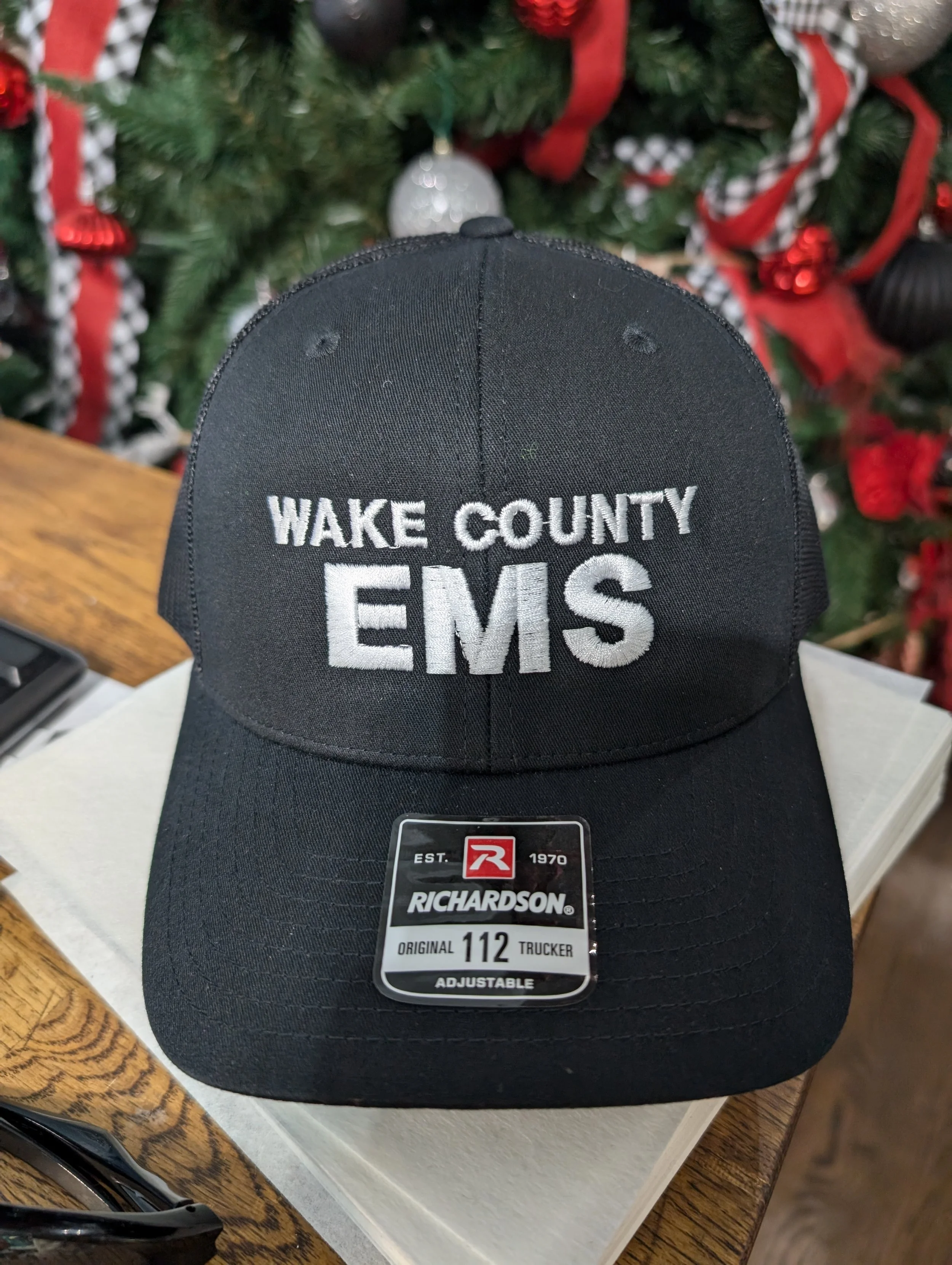 Black baseball cap with white embroidery that reads "WAKE COUNTY EMS". The cap is placed on a white surface with a Christmas tree in the background.
