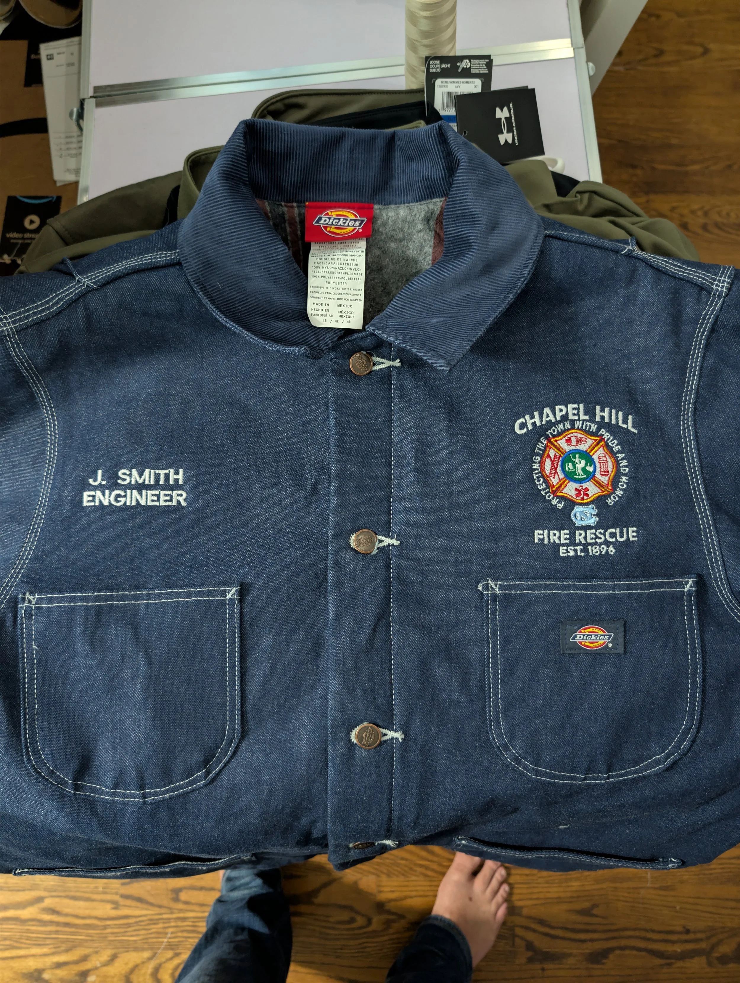 A dark blue work shirt with embroidered patches, including 'J. Smith Engineer' and 'Chapel Hill Fire Rescue,' with a Dickies logo on the front pocket, laid out on a surface with various items around it.