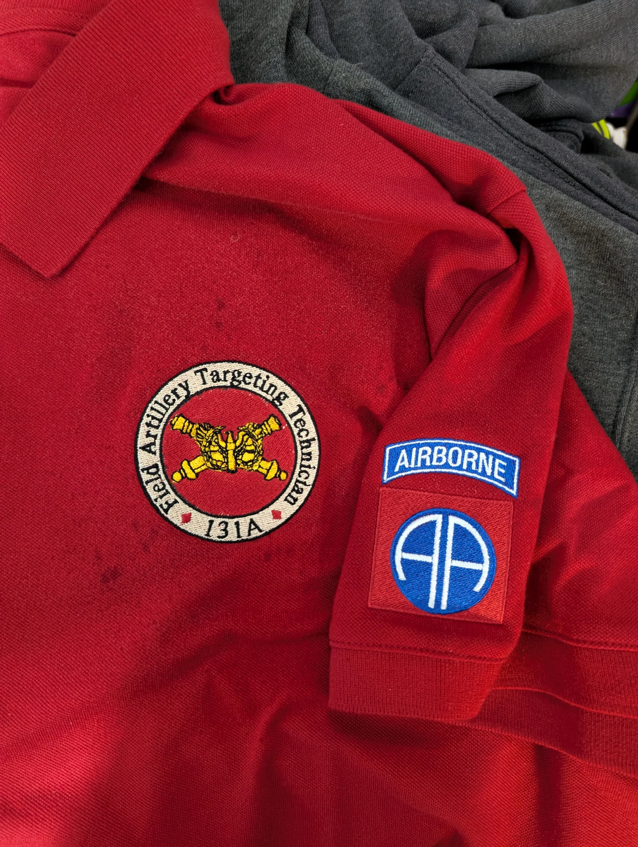 Close-up of a red uniform shirt with embroidered patches. One patch reads 'Field Artillery Targeting Technologies 131A' with an image of crossed cannons. The other patch is blue with 'AIRBOURNE' and a circular symbol.