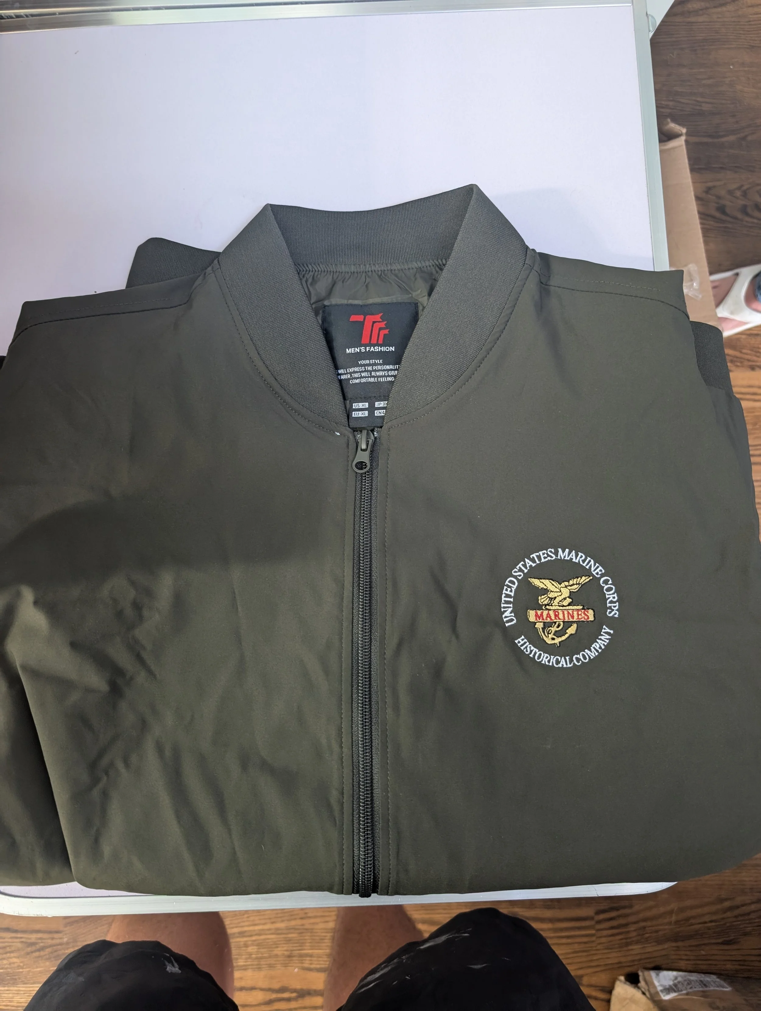 Olive green United States Marine Corps jacket with a Marine emblem and text embroidered on the front, lying on a white surface.