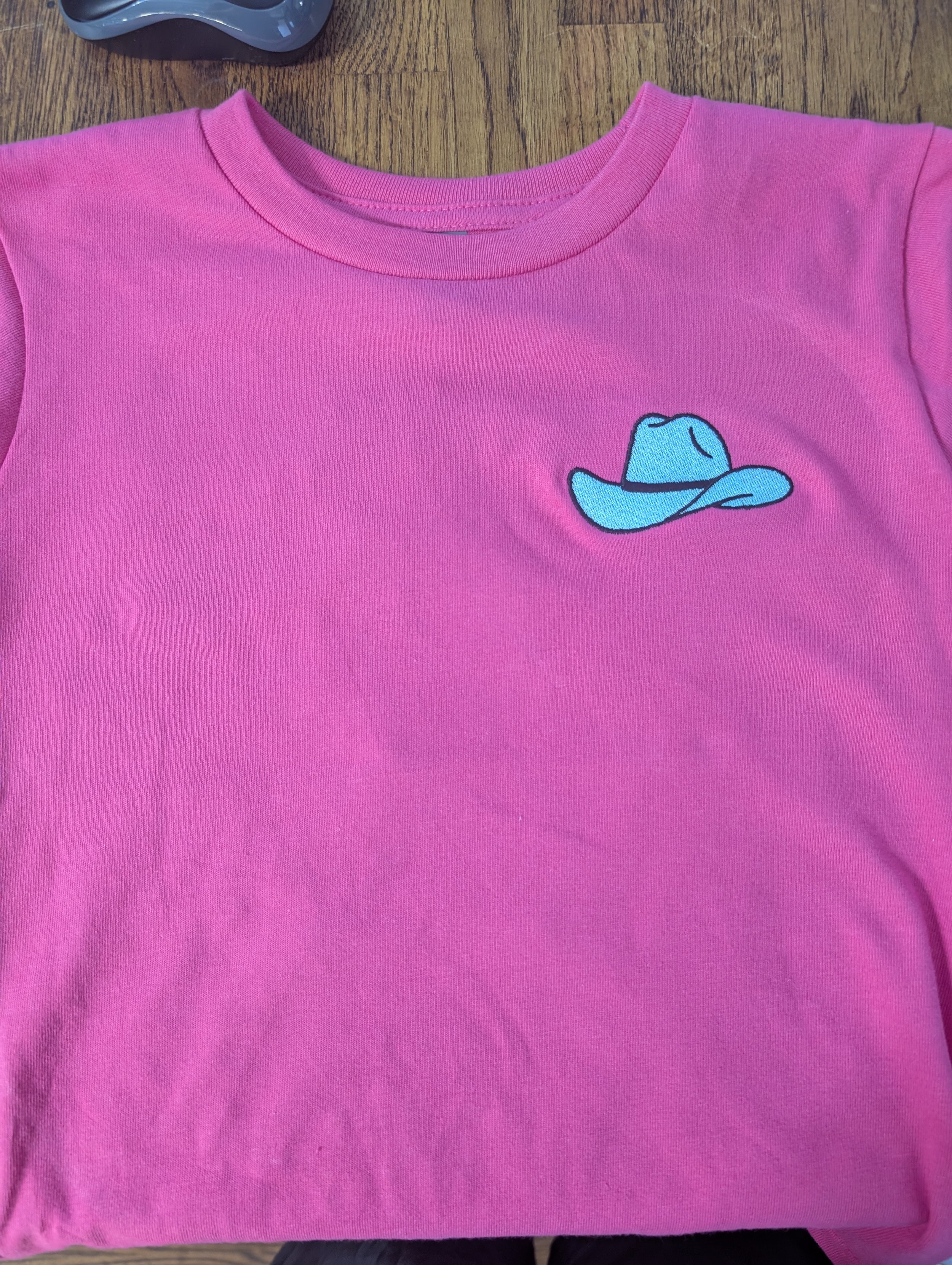 Pink shirt with a small embroidered blue cowboy hat design on the front left chest, lying on a wooden surface.