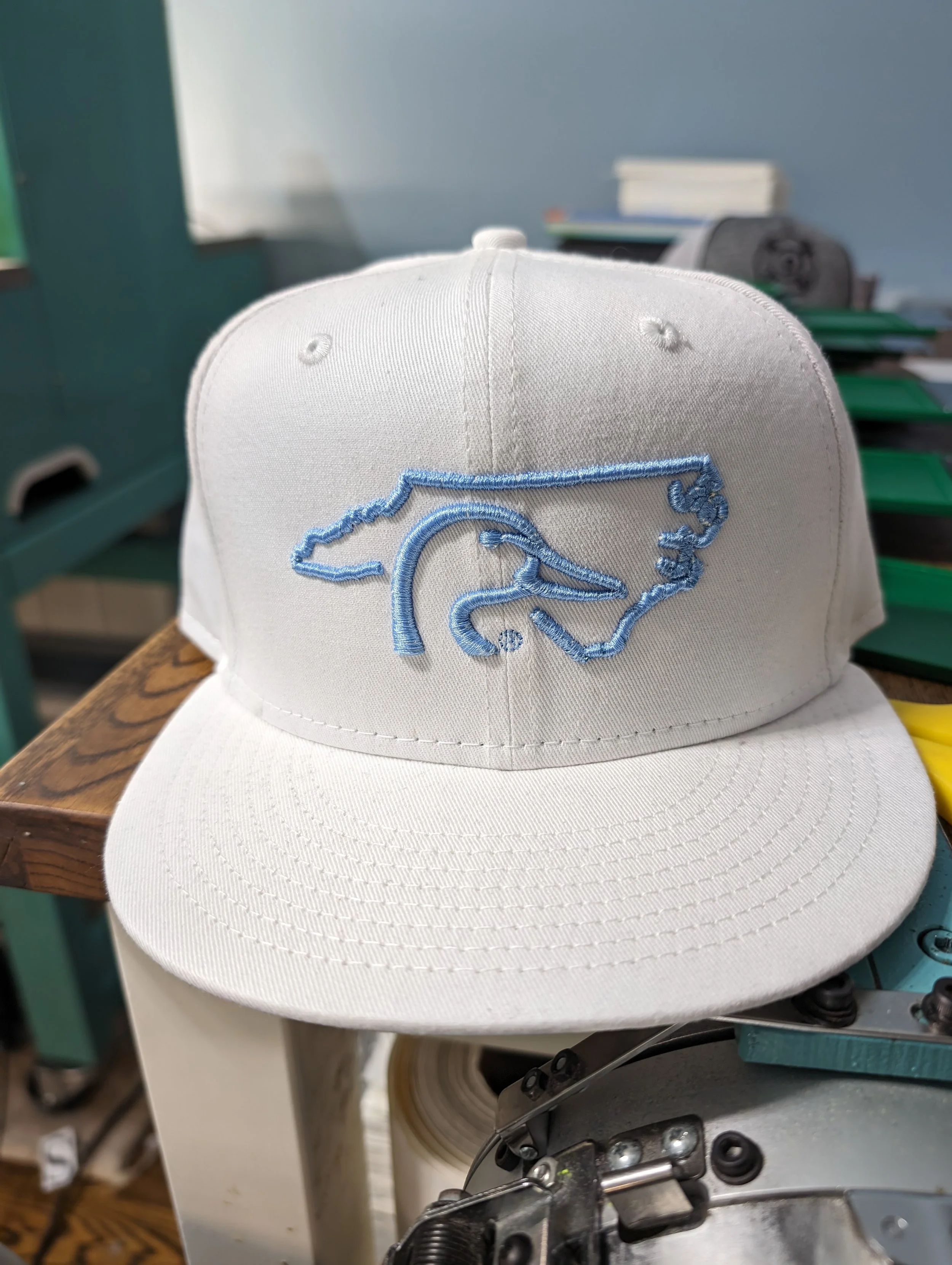 White baseball cap with a blue embroidered outline of the state of Florida and a stylized alligator logo on the front, placed on a wooden surface in a workshop.