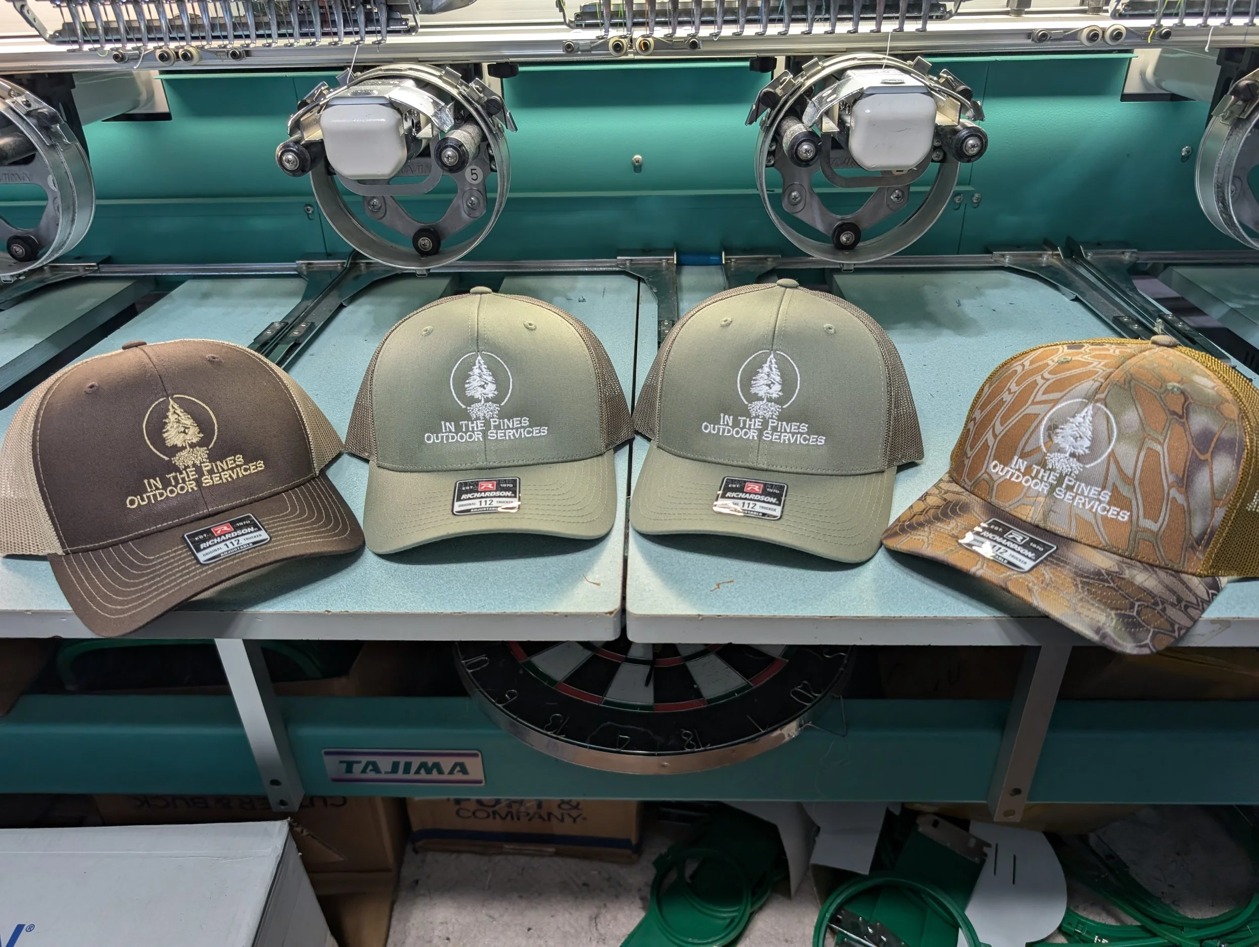 Four trucker caps with embroidery of a tree inside a circle and the text "In the Pines Outdoor Services" on a blue workbench, with parts of a machinery or equipment above and a dartboard below.