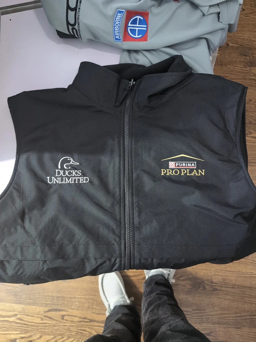 Black vest with white and gold logos reading 'Ducks Unlimited' and 'Purina Pro Plan' laid flat on a surface, with a person wearing white shoes and dark pants standing below.