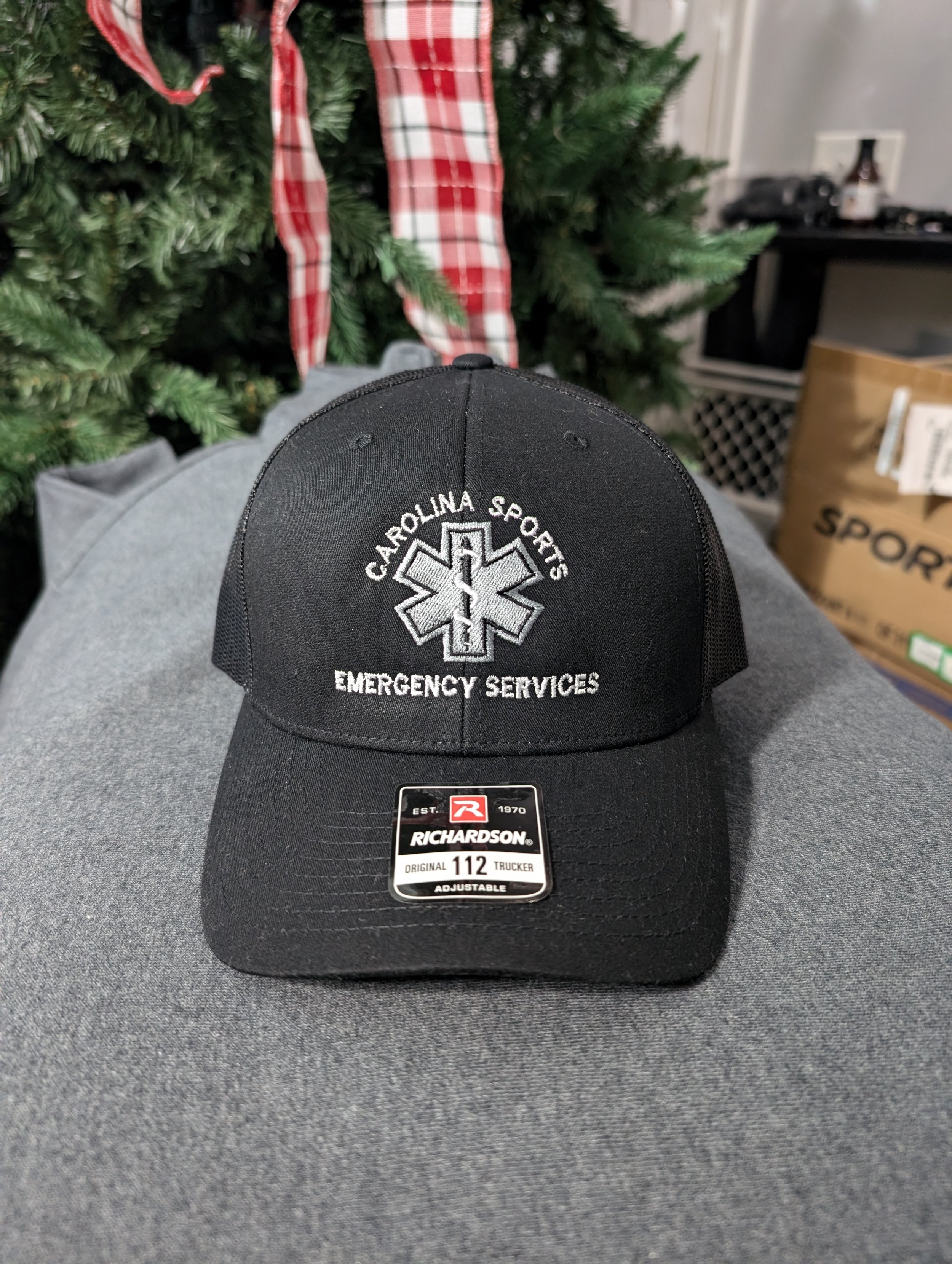 A black baseball cap with embroidery for Carolina Sports Emergency Services and a medical symbol, placed on a person's lap with a Christmas tree and set-up in the background.