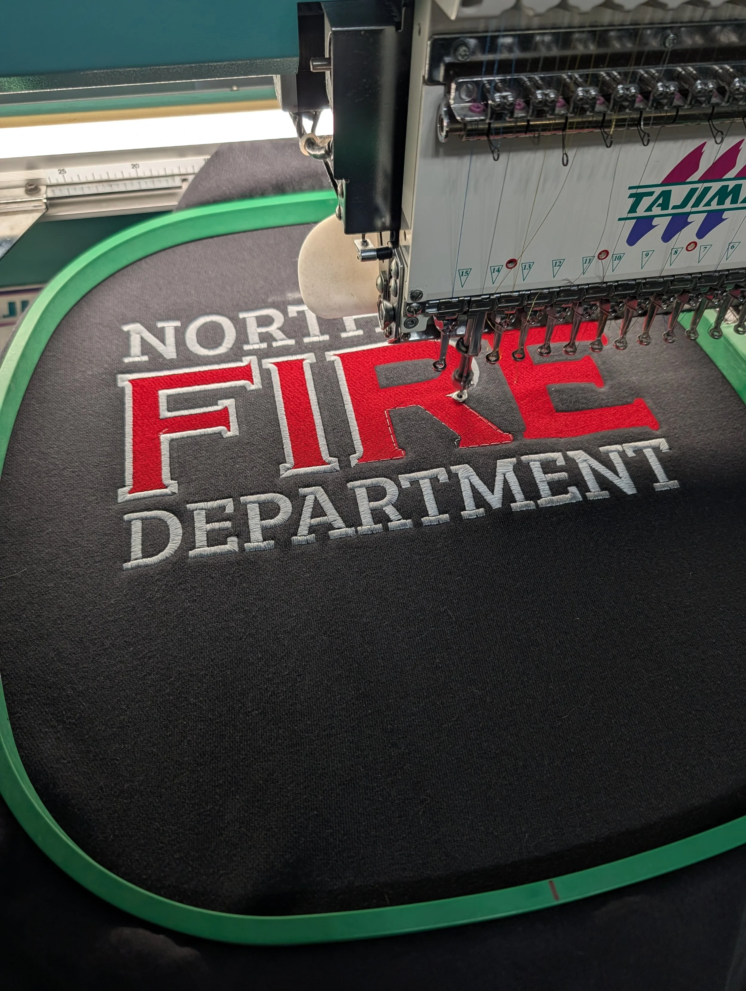 Embroidery machine stitching the words 'North Fire Department' onto black fabric.