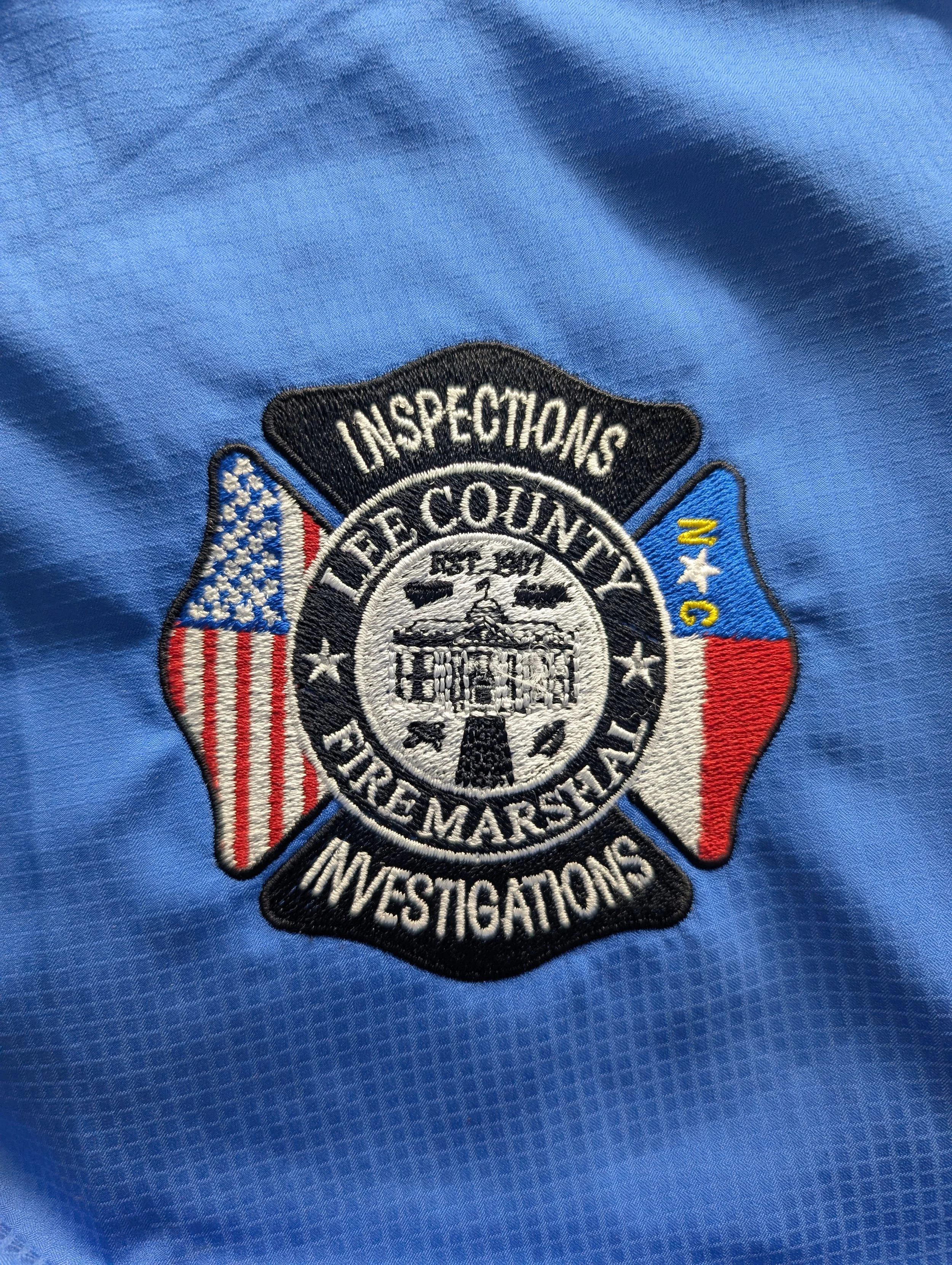 Close-up of a light blue uniform shirt with an embroidered badge for Lee County Sheriff's Office Investigations, featuring the American flag and Texas flag, with the text "Inspections" and "Investigations".