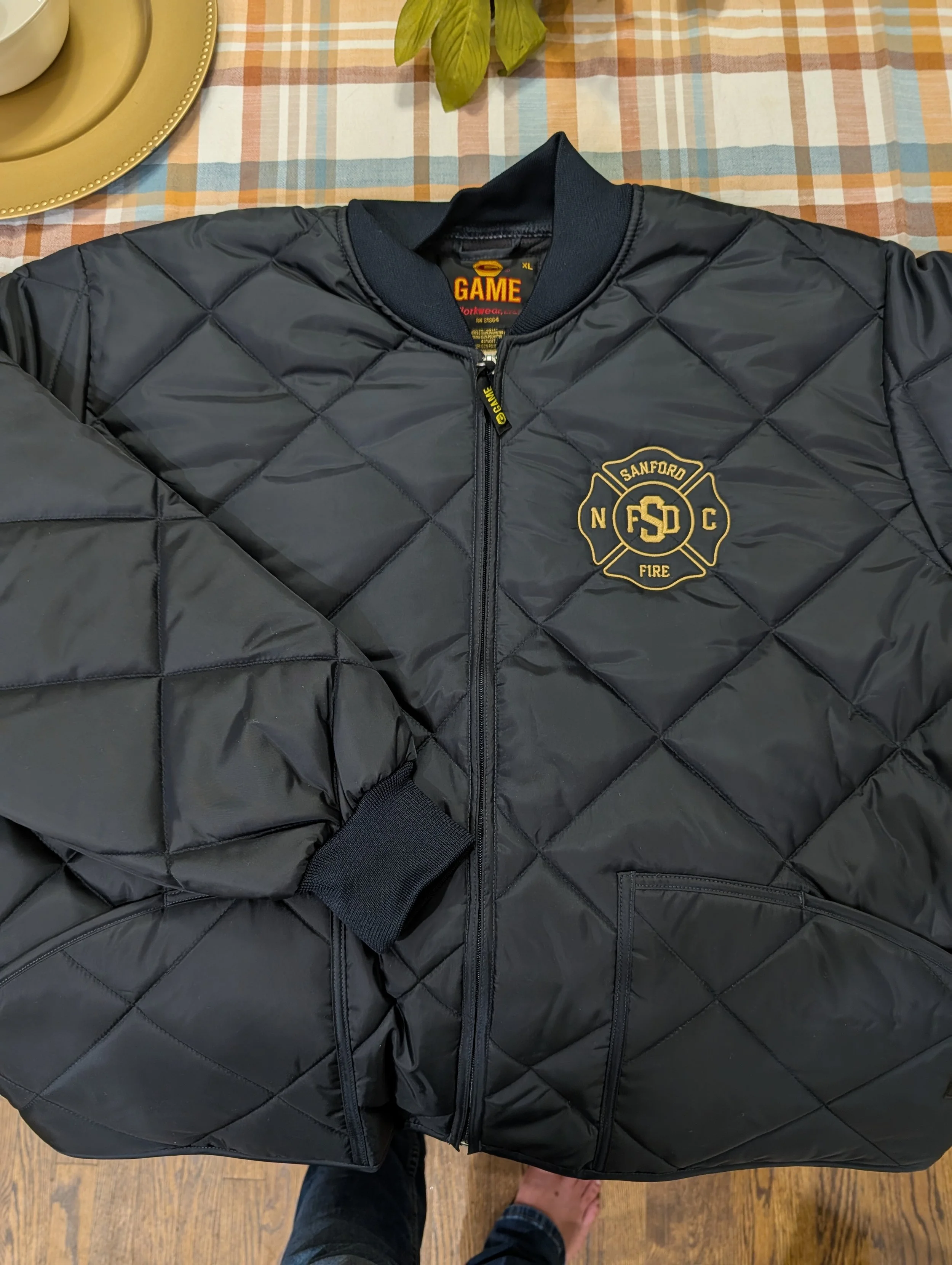 Black quilted jacket with Fire Department logo on the front, lying on a table with a plaid tablecloth.