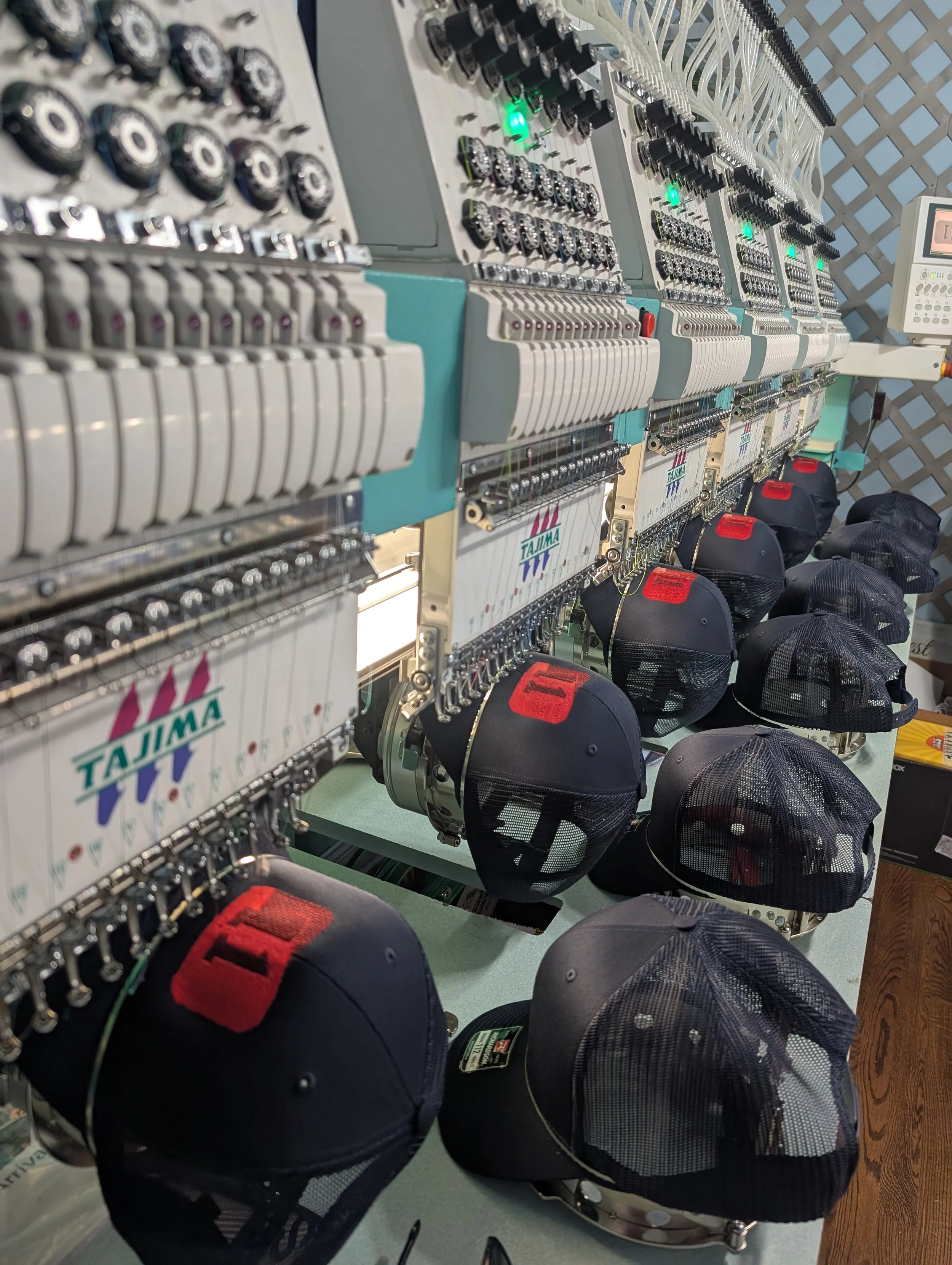 Embroidery machine sewing baseball caps with black mesh and gray fabric, some with red patches, on a green table in a workshop.