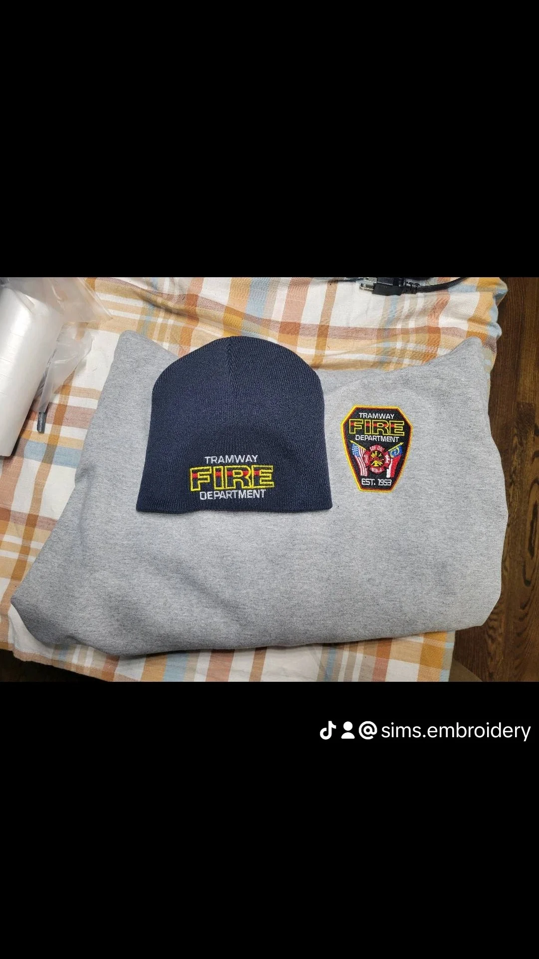 A gray sweatshirt with two embroidered patches: one with the text "TRAMWAY FIRE DEPARTMENT" and a firefighter badge, the other with the same text and logo on a black background, a navy blue knit beanie with "TRAMWAY FIRE DEPARTMENT" embroidered in ye