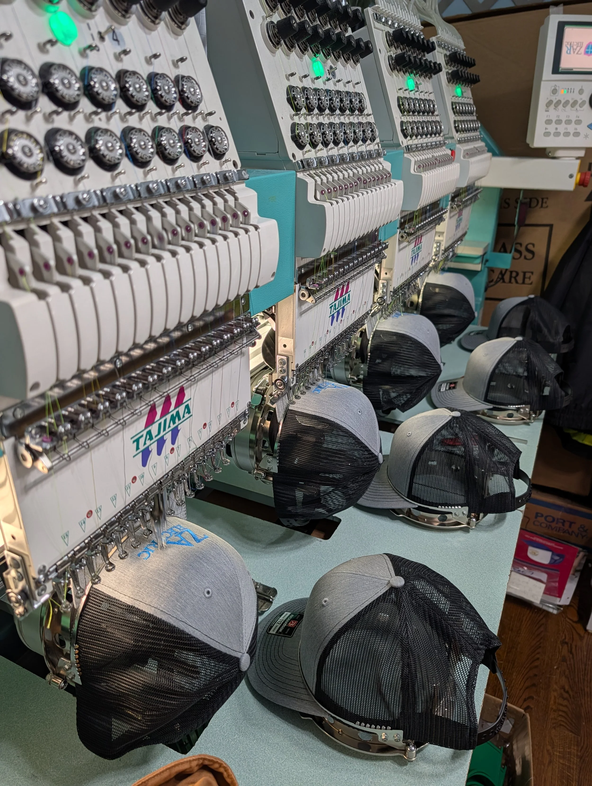 Multiple embroidery machines set up on a table with several baseball caps in front of them, ready for embroidery. The caps are gray with black mesh, and the machines are in operation, with threads visible.