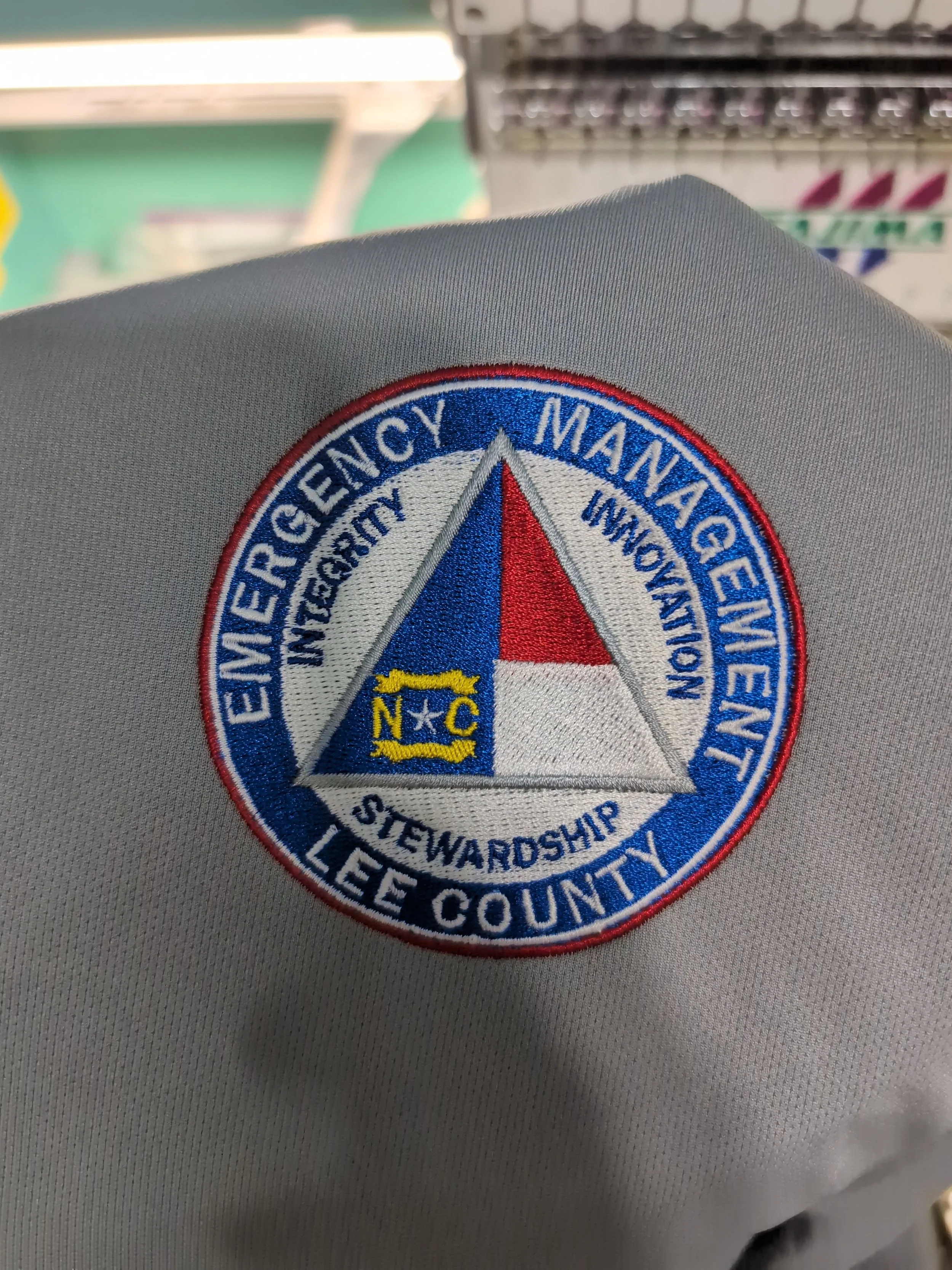 Close-up of an embroidered patch on gray fabric, featuring a blue circle with text around the border reading 'Emergency Management,' 'Stewardship,' 'Lee County,' and other words. The center of the patch depicts a triangle with red, white, and blue se