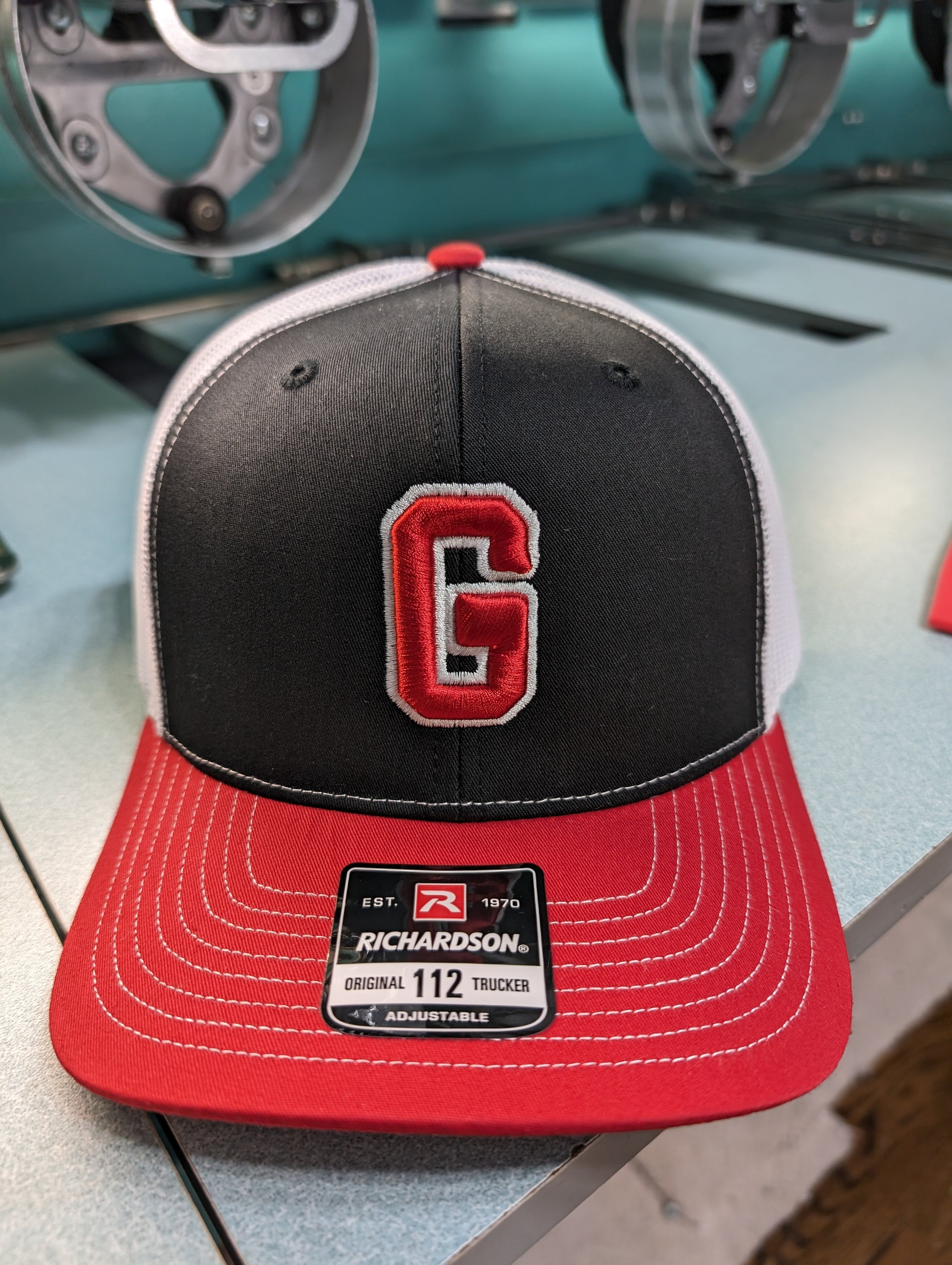 A baseball cap with a black front panel featuring a red outlined letter G. The cap has a red brim with white stitching and a mesh back with a white border.