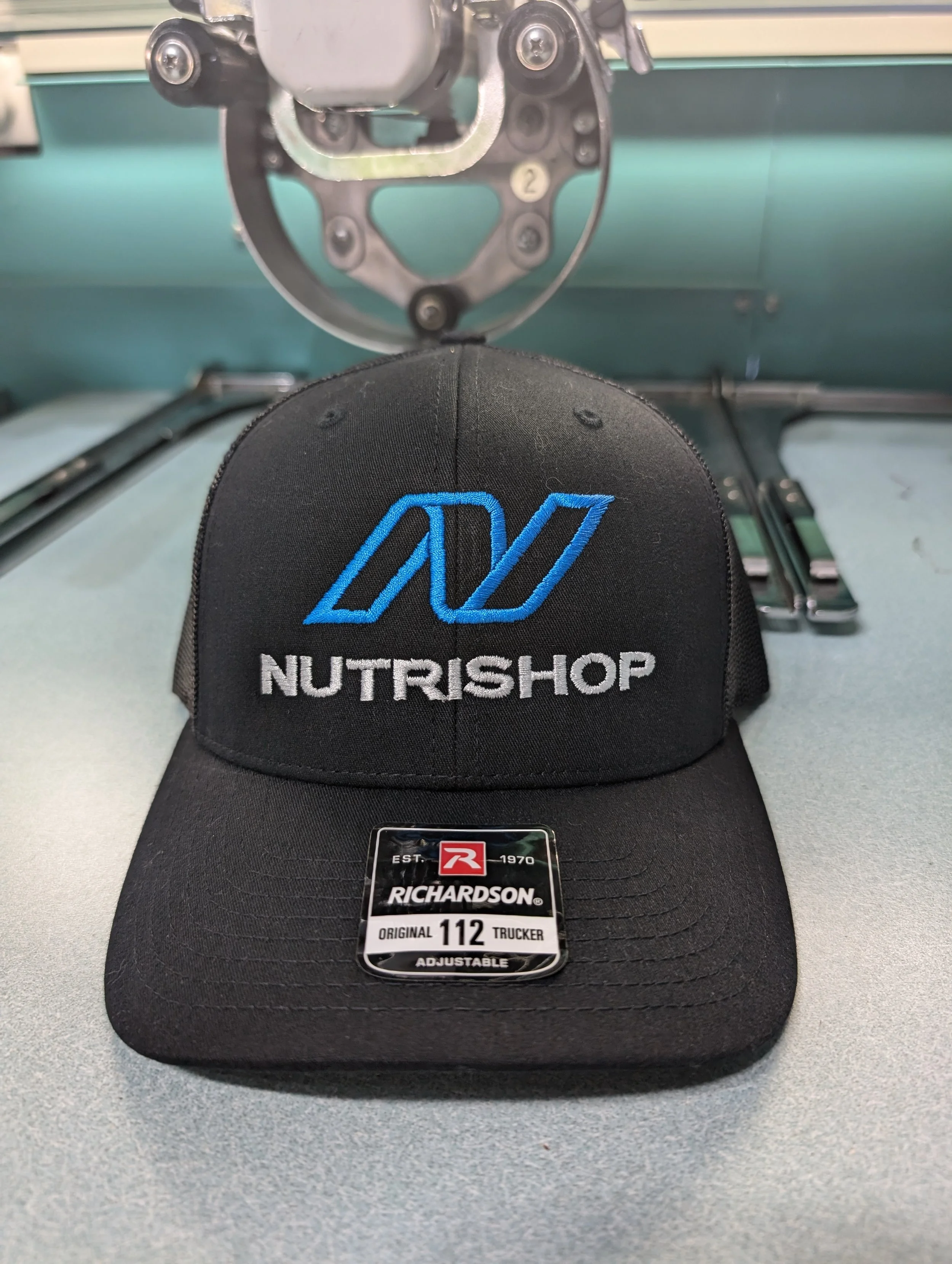 Black baseball cap with blue and white embroidery of NutriShop logo and text, placed on a desk in front of machinery.