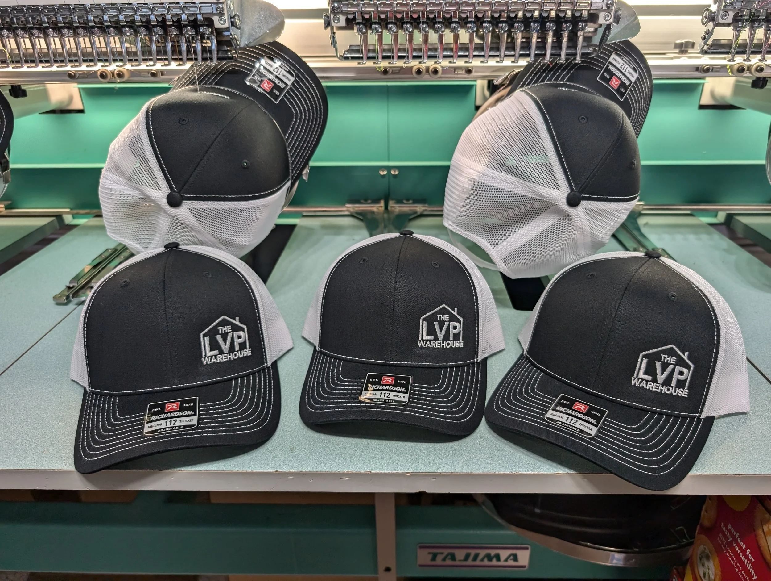 Six black and white trucker hats with the logo 'The LVP Warehouse' displayed on a green workbench, with three hats lined up in front and three hats hanging over hooks above.