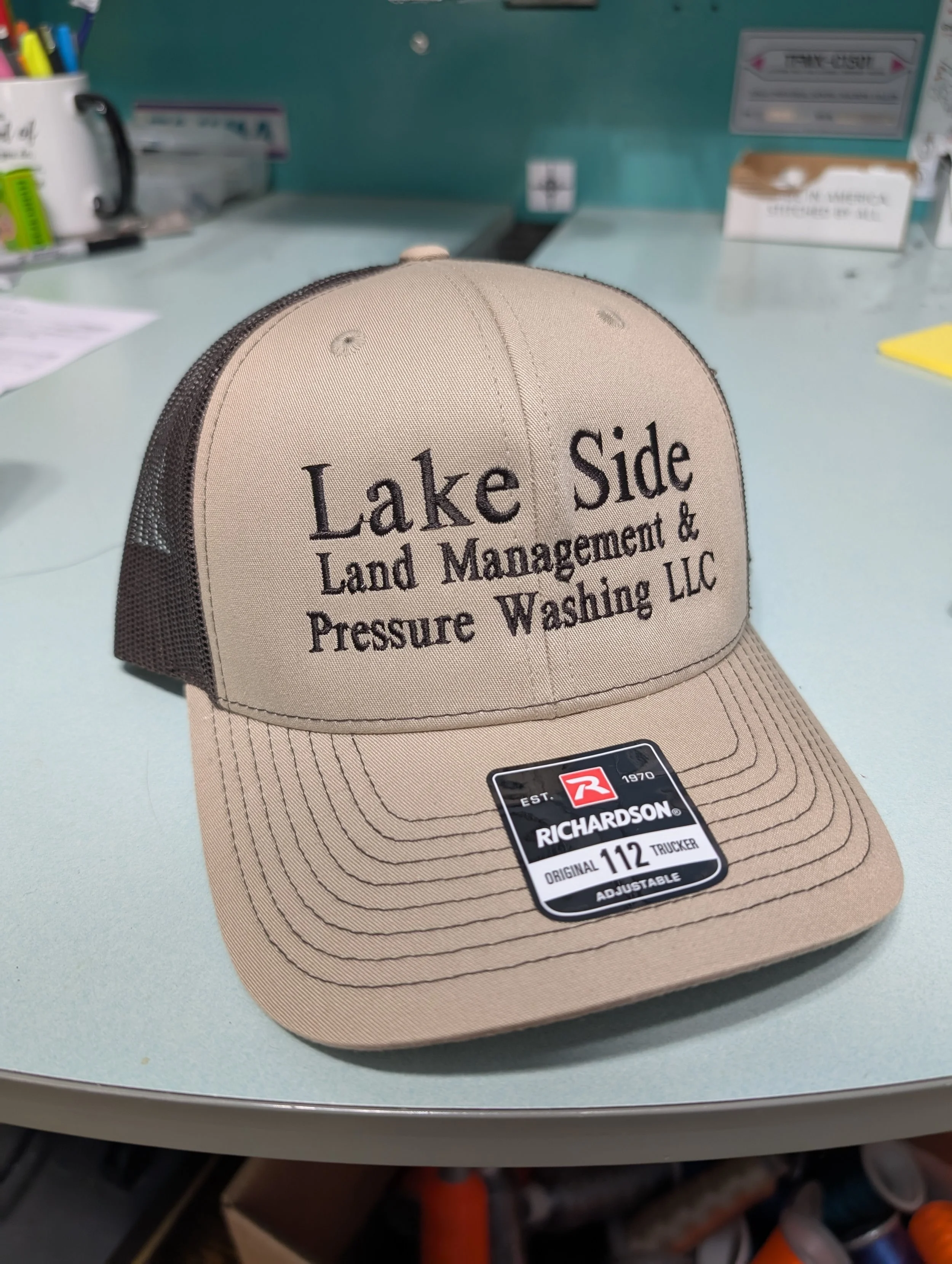 Tan baseball cap with mesh back and embroidered text reading 'Lake Side Land Management & Pressure Washing LLC' on the front, placed on a light-colored table.