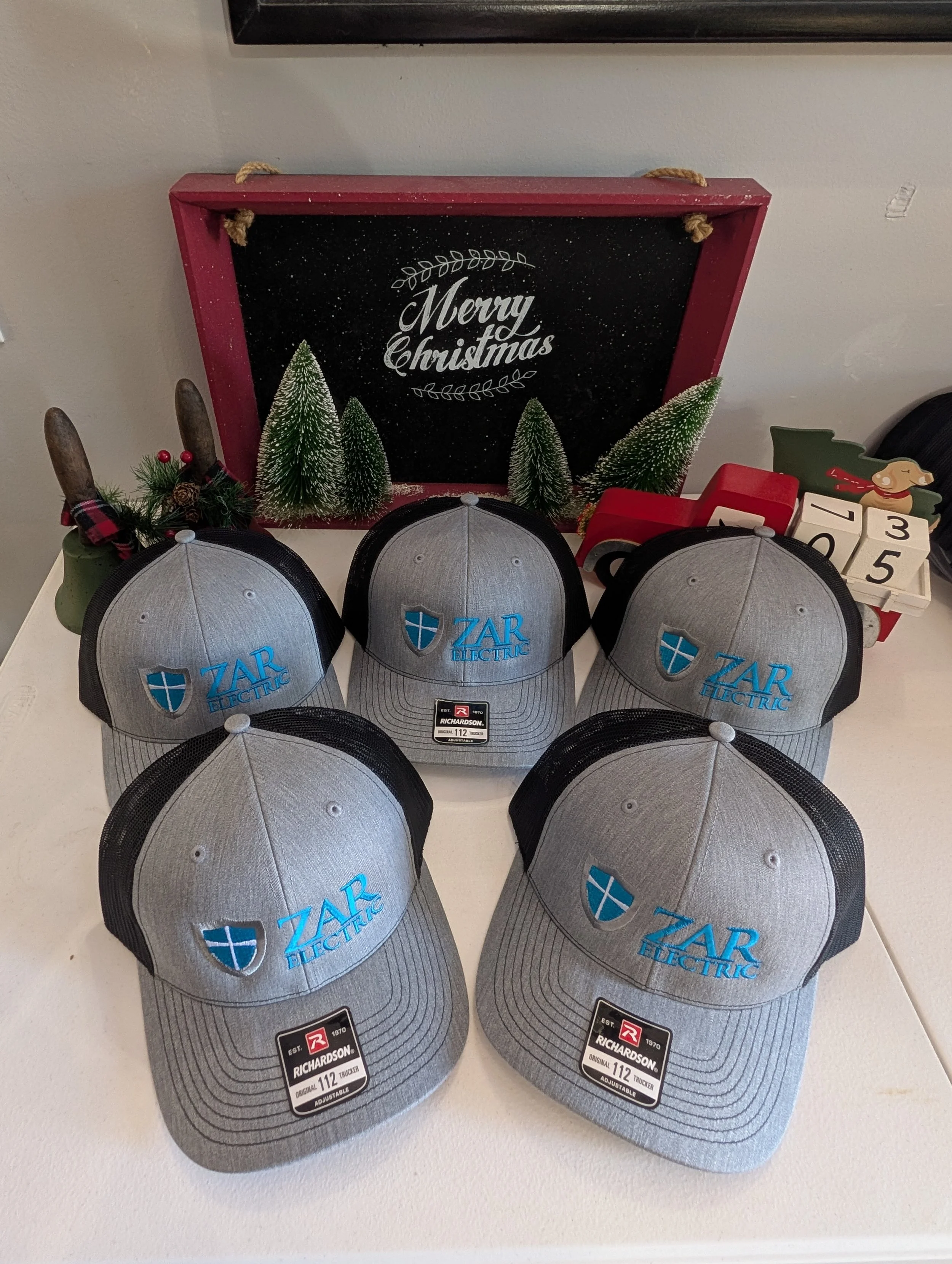 Six gray and black ZAR Electric trucker hats with blue logo, arranged in two rows, on a table with Christmas decorations and a Merry Christmas sign in the background.