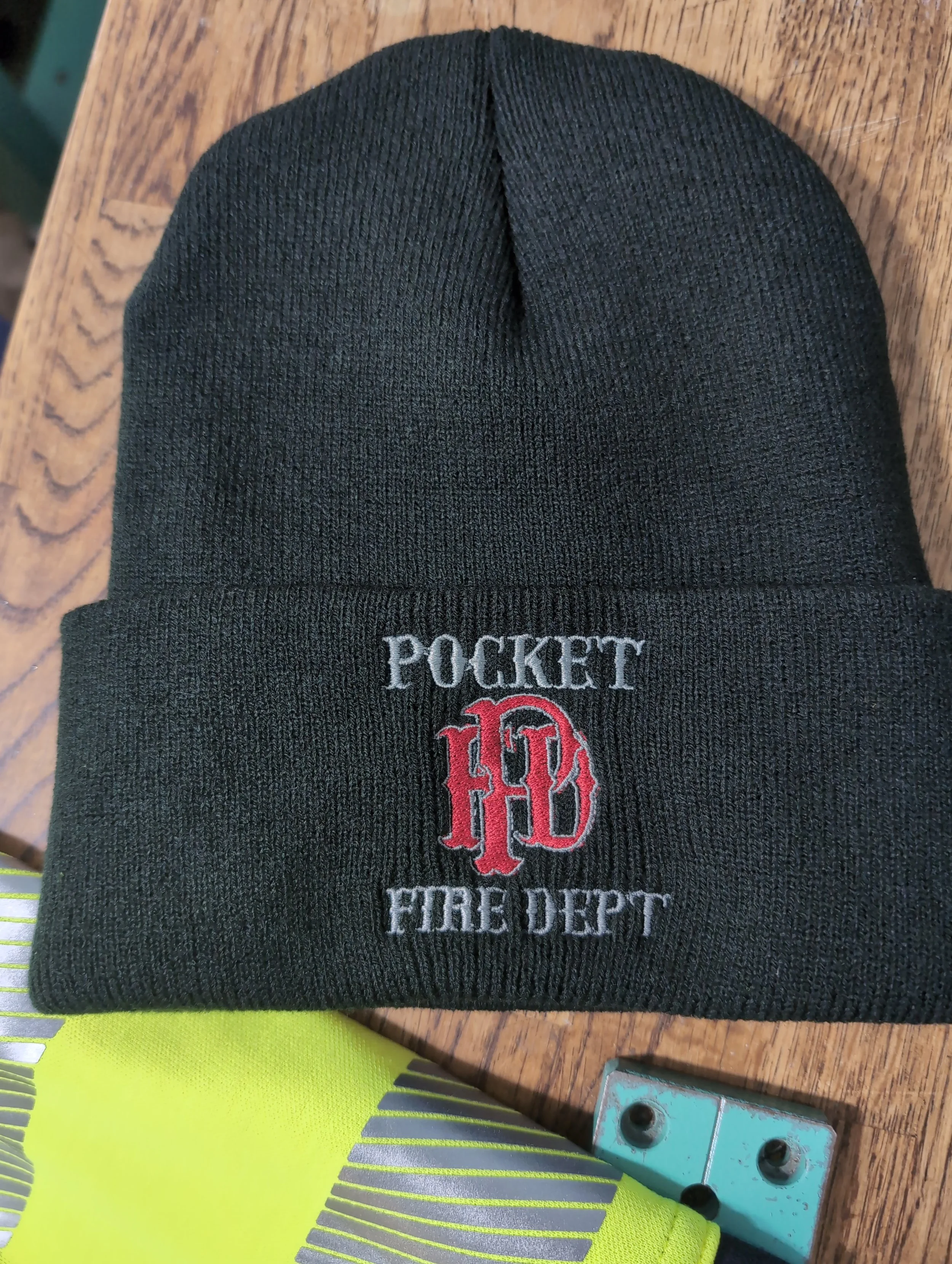Black knit beanie with an embroidered patch reading 'Pocket Fire Dept' and a red firefighter emblem on the front, placed on a wooden surface.