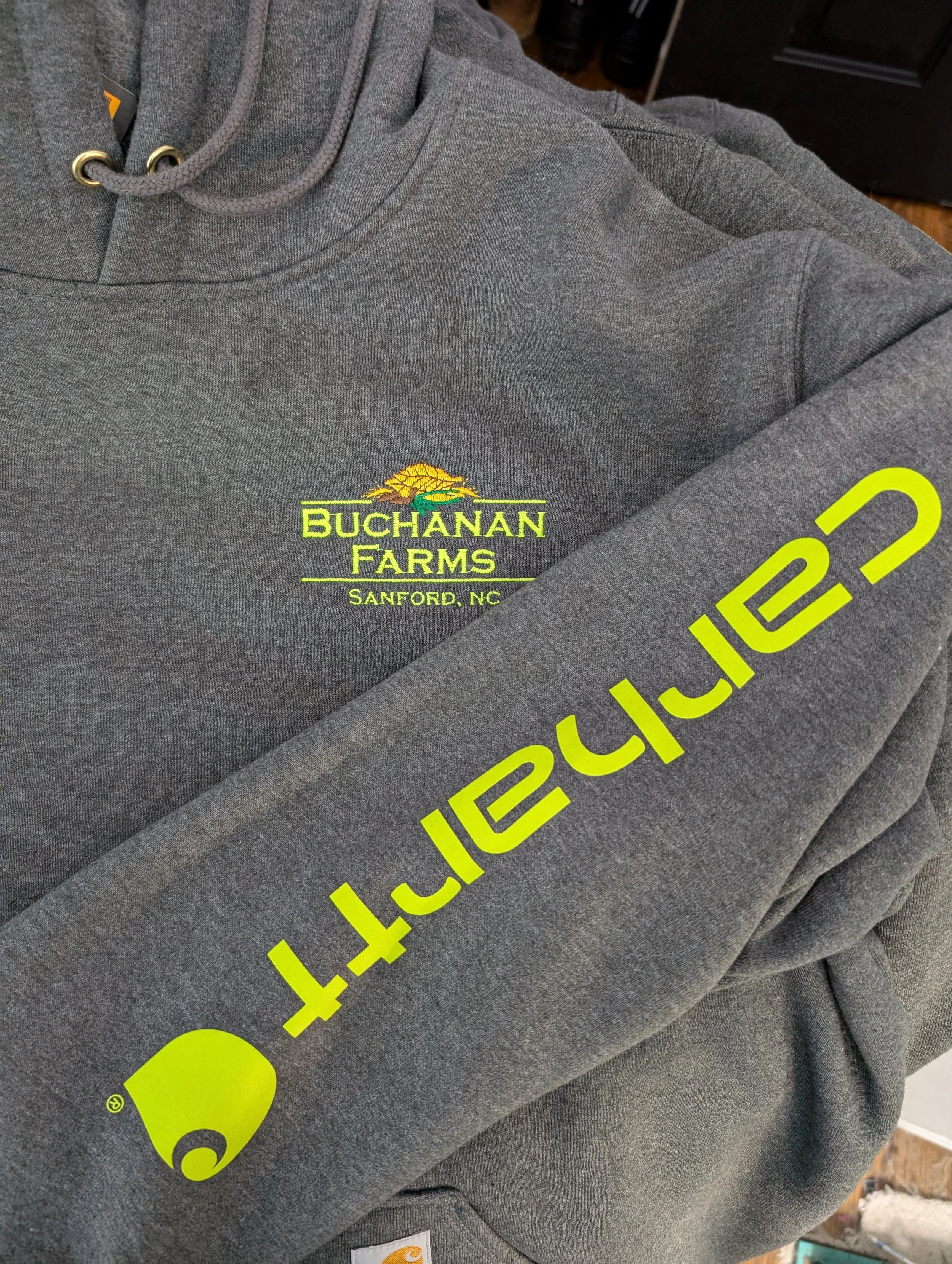 Close-up of a gray Carhartt sweatshirt with neon yellow and green embroidery that reads "Buchanan Farms, Sanford, NC" and a neon yellow Carhartt logo on the sleeve.