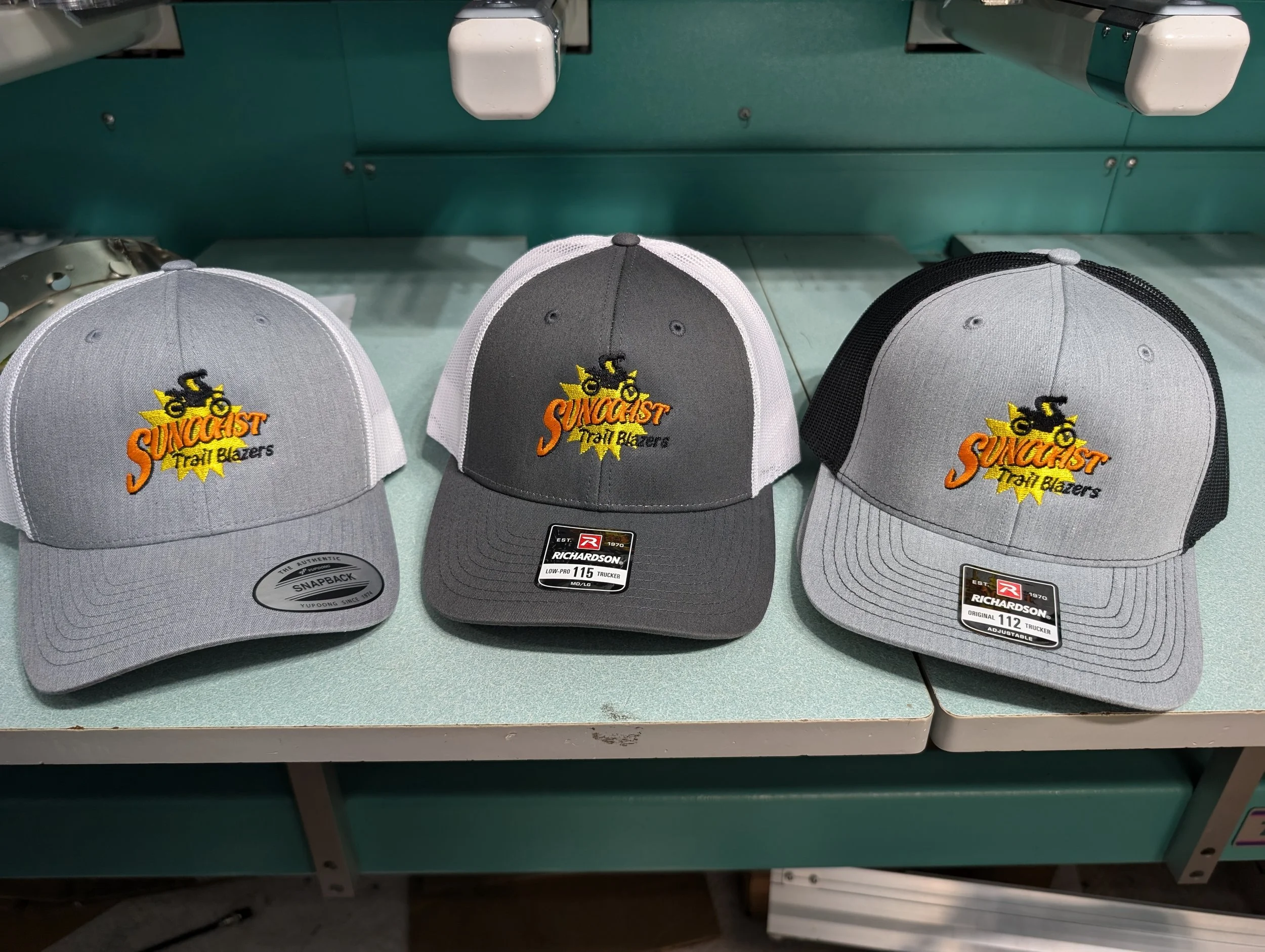 Three baseball caps with the "Suncoast Trail Blazers" logo, featuring a bike graphic and yellow starburst, displayed on a table with a teal background.