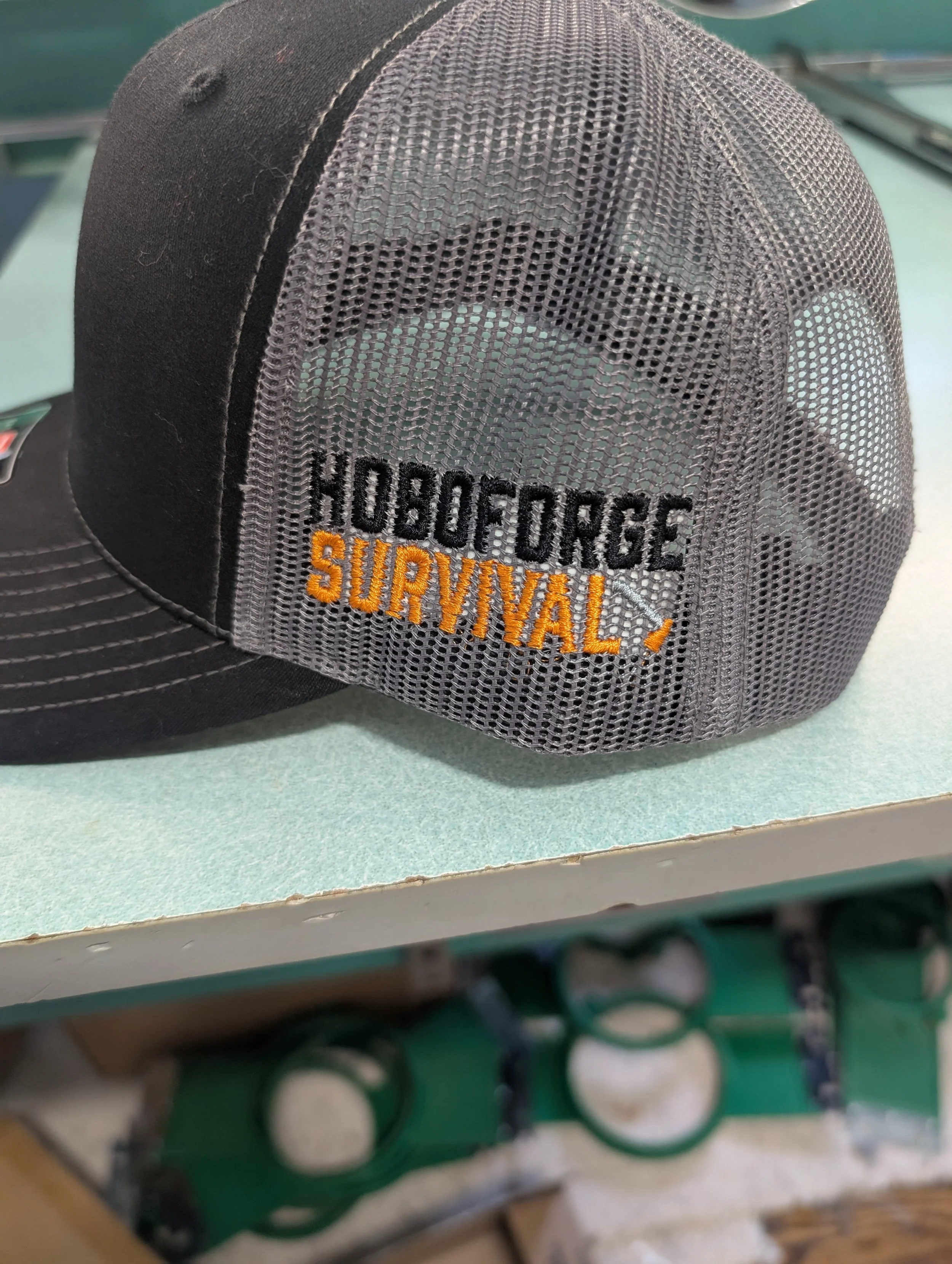 Black trucker hat with mesh back panel that has the embroidered words "HoboForge Survival!" in black and orange.