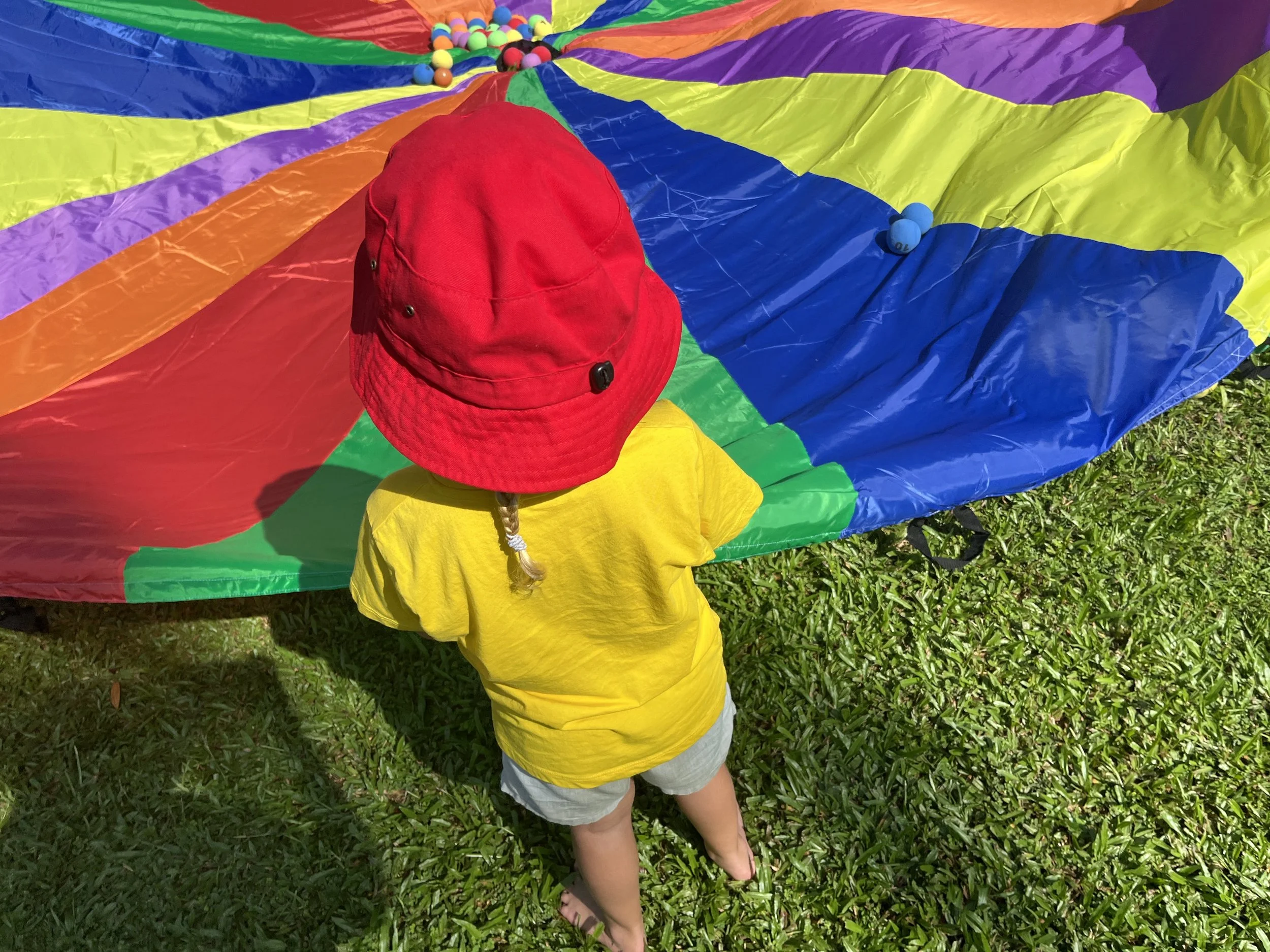 Child with a red hat and yellow t-shirt standing on grass, holding a rainbow-colored parachute with small balls on top.