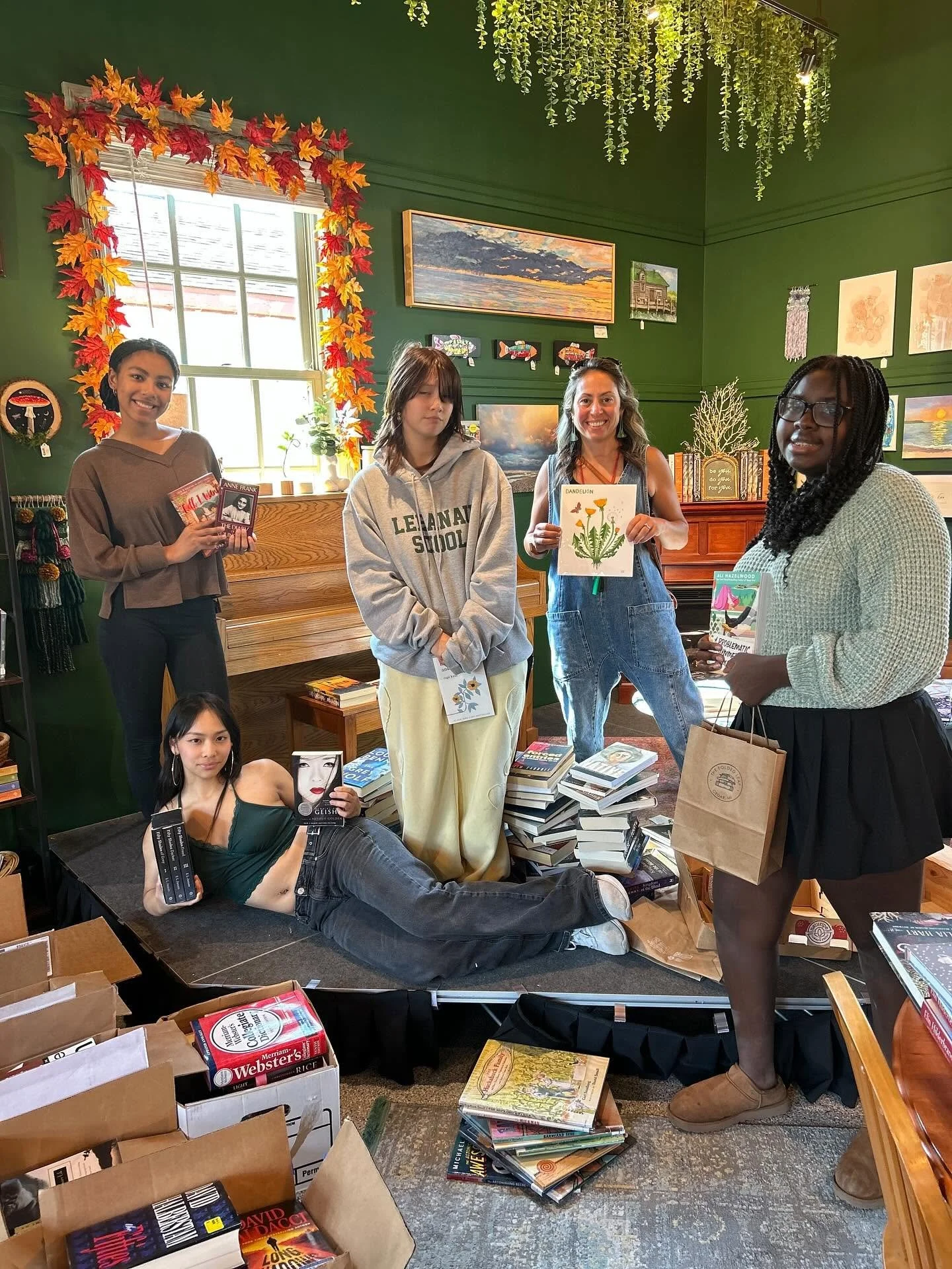 Five women and one girl posing in a bookstore with books, artwork, and autumn decorations in the background. One woman is lying on the floor holding books, while others hold books and a drawing of a dandelion.