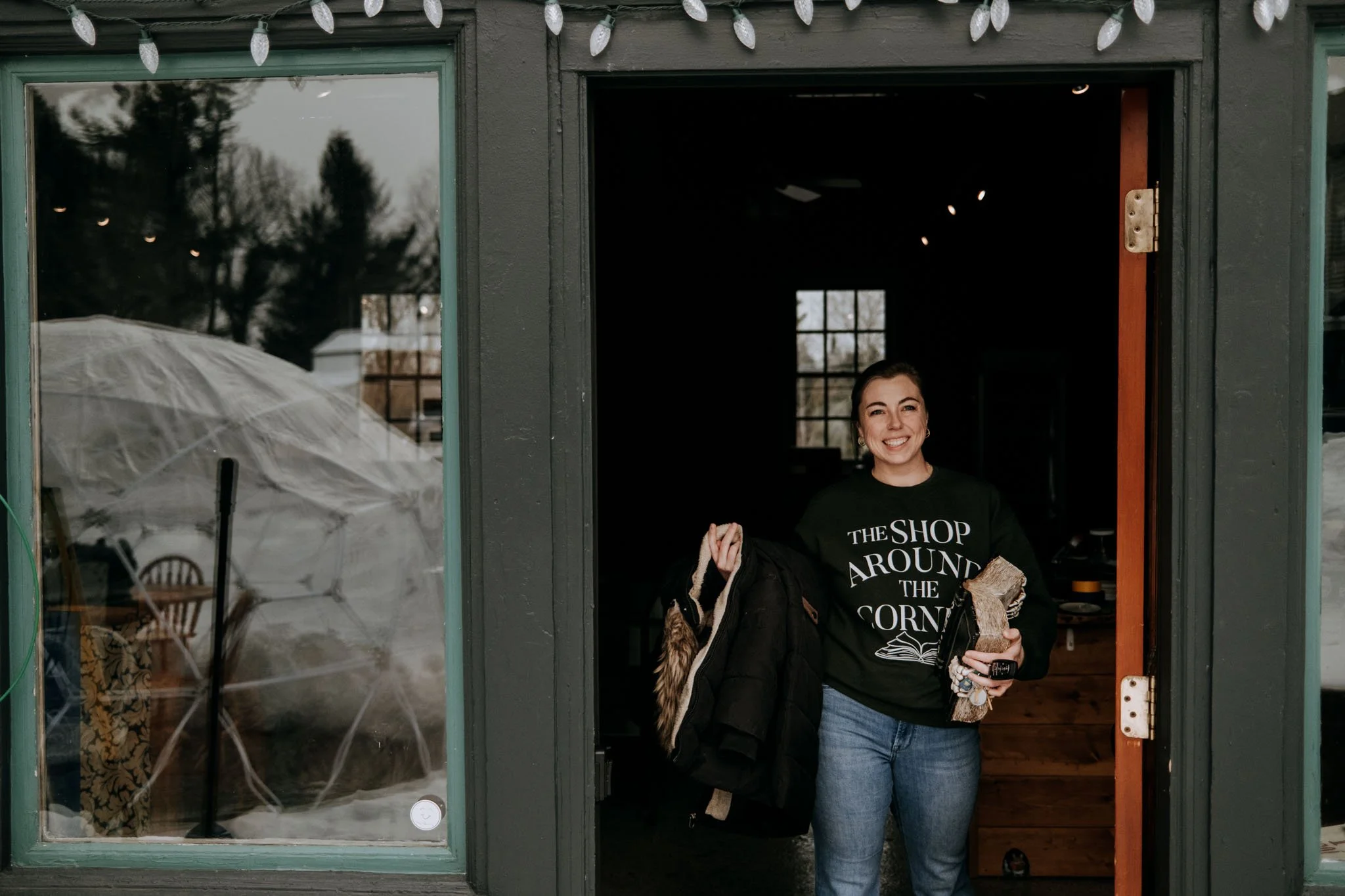 A woman smiling standing in the doorway of a building, holding a coat and a bag. She is wearing a black sweatshirt with white text that reads 'The Shop Around The Corner' and blue jeans. The building has dark green trim, with string lights hanging above.