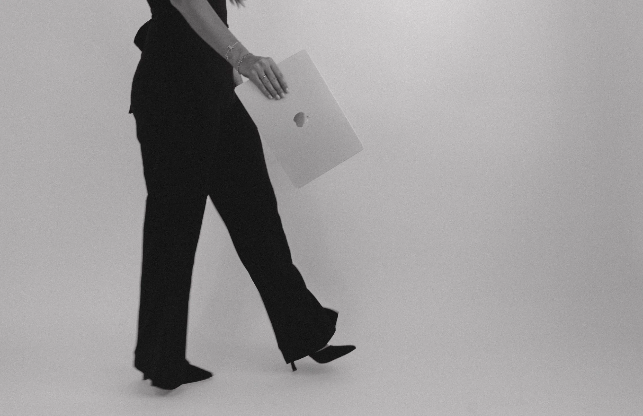 A person in business attire holding a closed laptop, walking against a plain background.