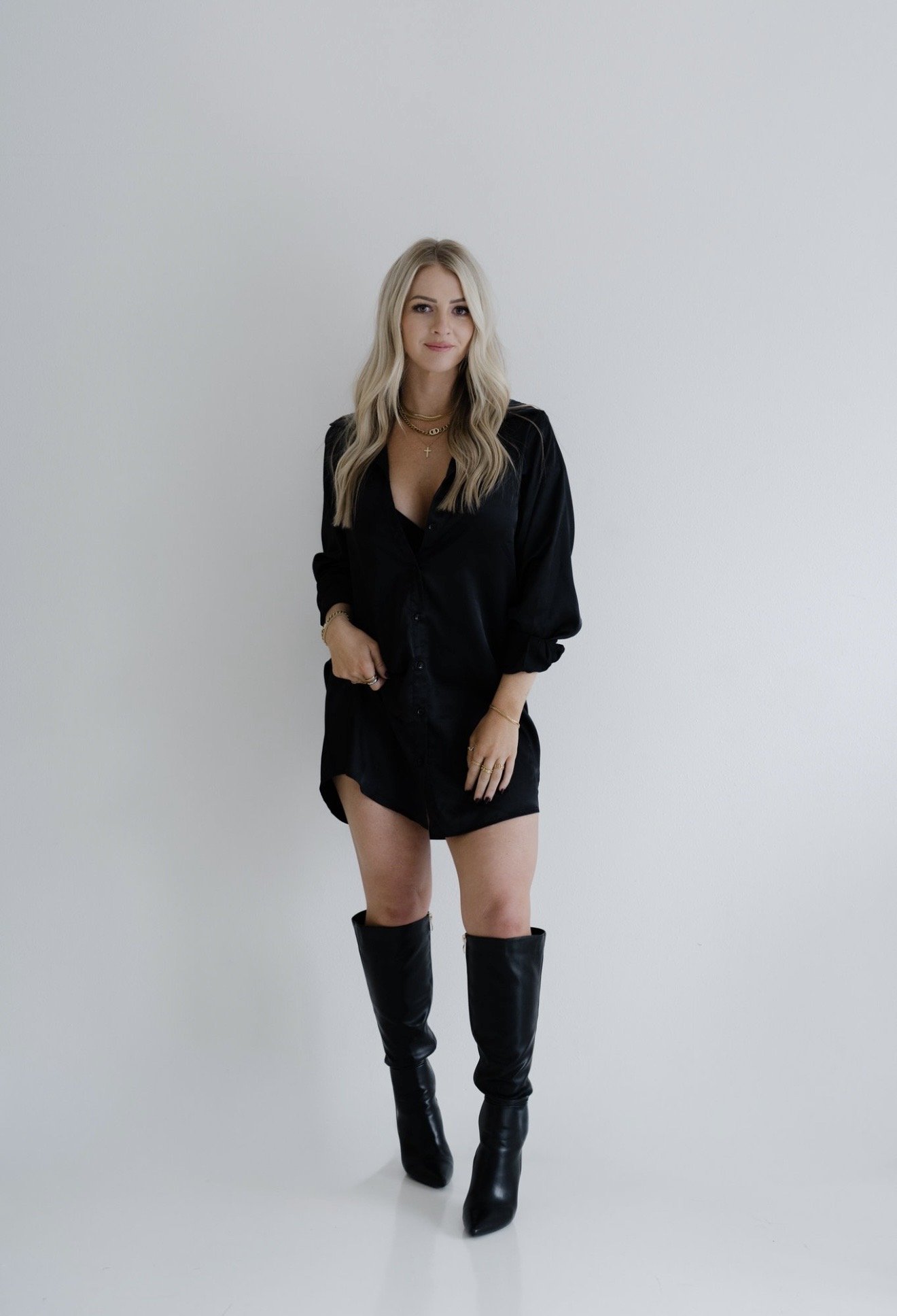 A woman with long blonde hair wearing a black shirt, black shorts, and tall black boots, standing against a plain white background.