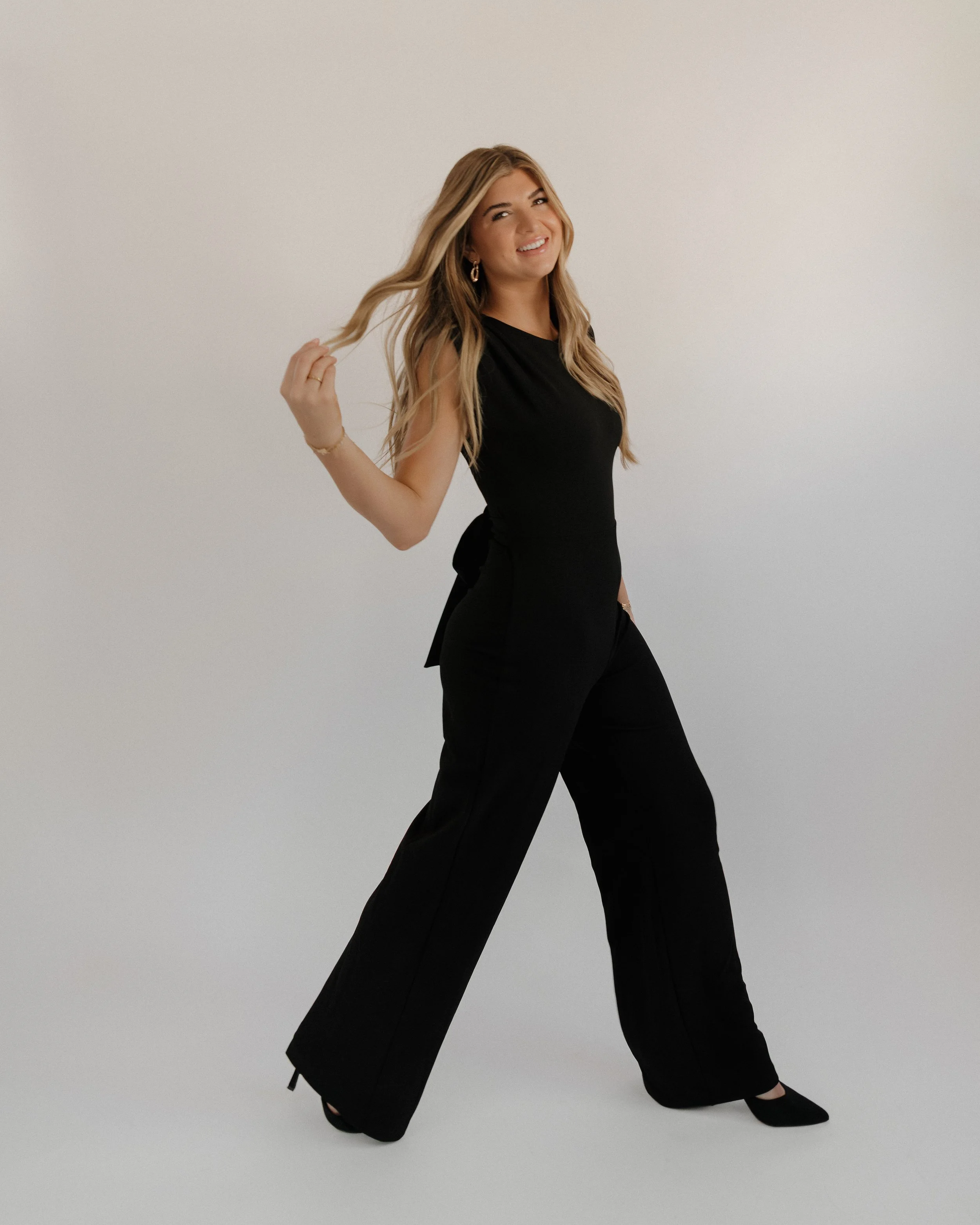 A woman with long blonde hair, wearing a black sleeveless top and black wide-leg pants, smiling and holding a strand of her hair in a studio with a plain light background.