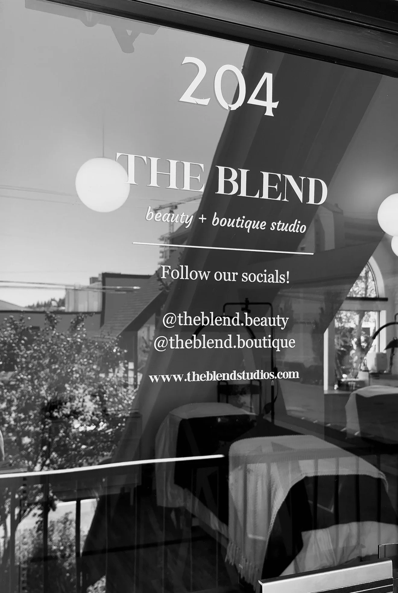 Glass window with information for THE BLEND beauty and boutique studio, including social media handles and website, with reflections of the street and furniture inside.